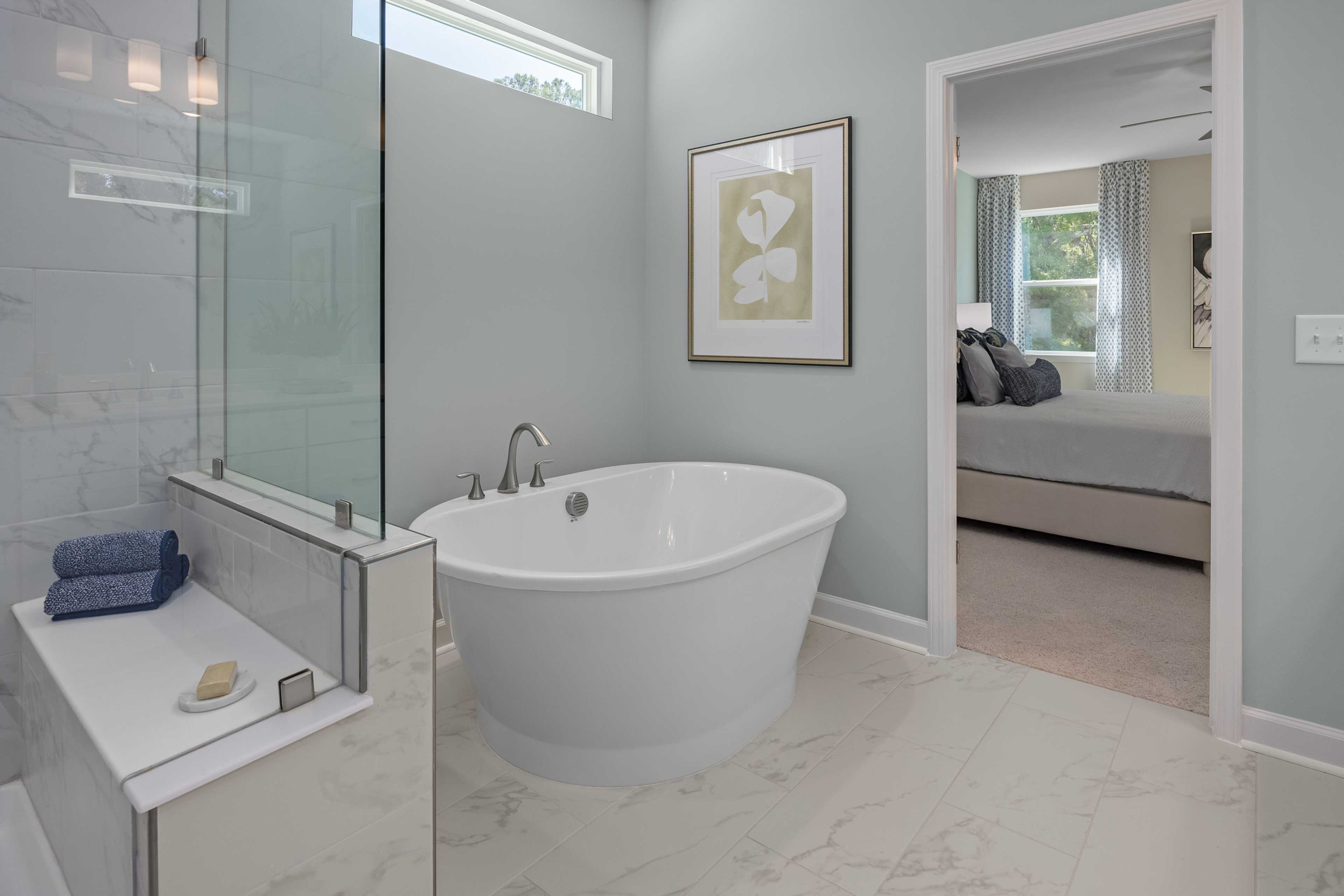 Spa-like primary bathroom at Sage on North Main in Wake Forest NC with freestanding white tub, glass shower, and bedroom view