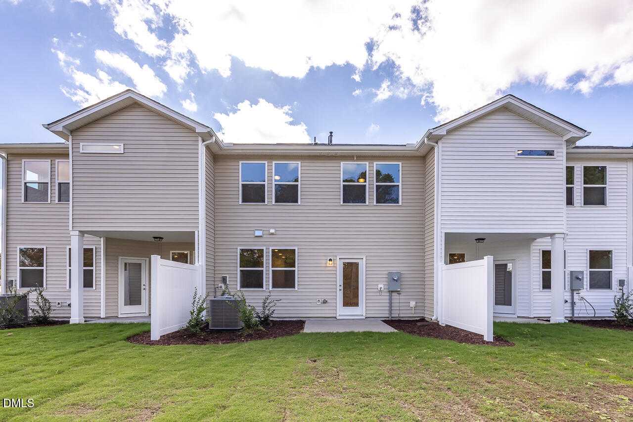 Rear view of two-story townhomes with beige siding, fenced patios, AC units, and green lawns in Springvale, Fuquay-Varina, NC