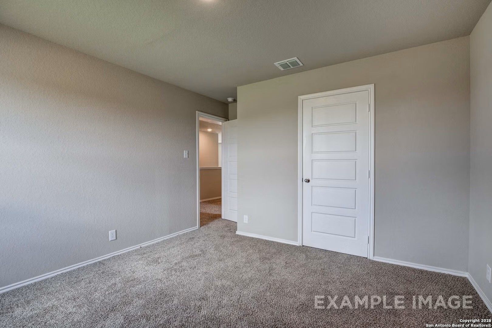 Bright bedroom with beige walls, carpet flooring, white door, and adjacent bathroom access in Davidson Homes The Murray K, San Antonio, Texas