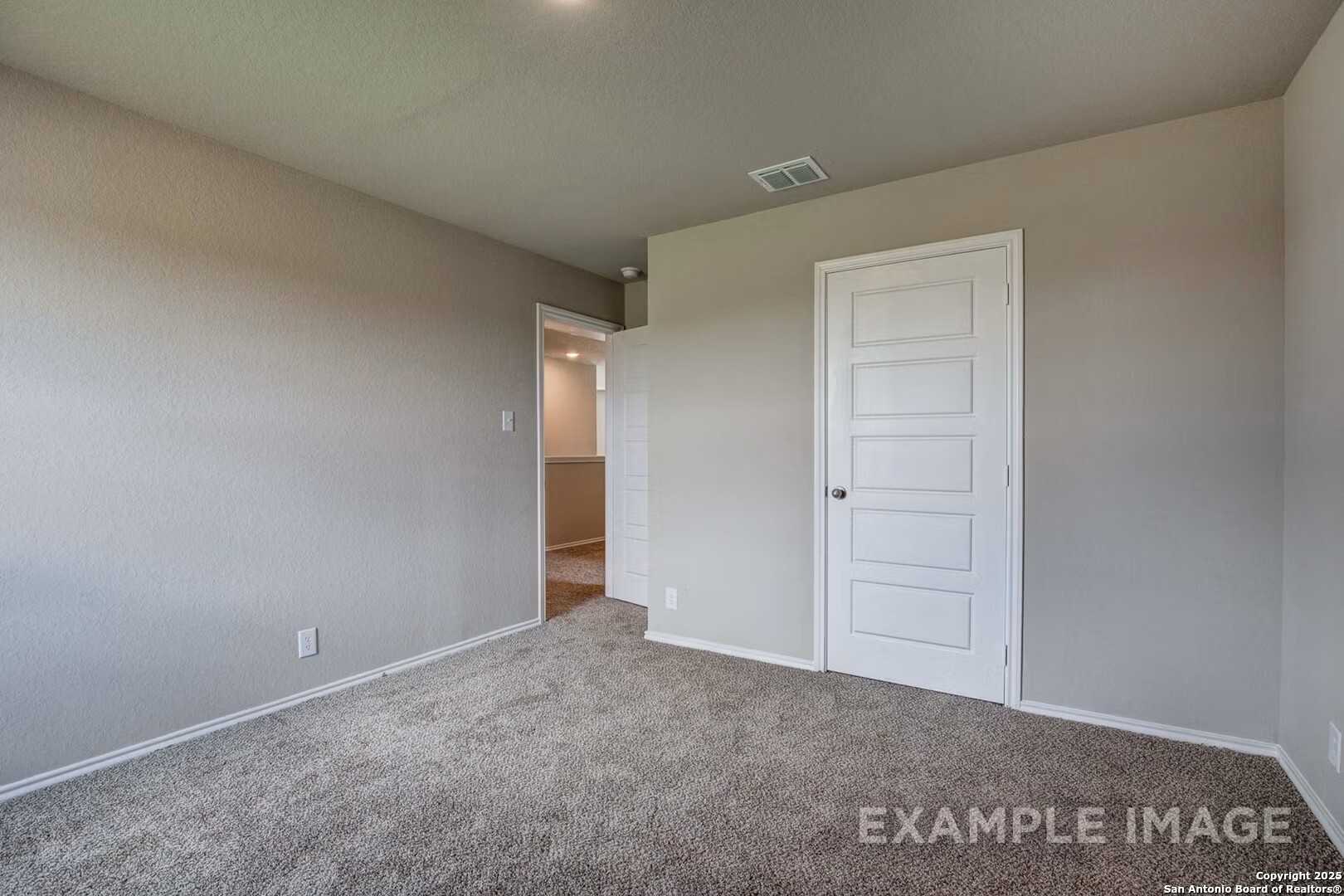 Bright bedroom with beige walls, carpet flooring, white door, and adjacent bathroom access in Davidson Homes The Murray K, San Antonio, Texas