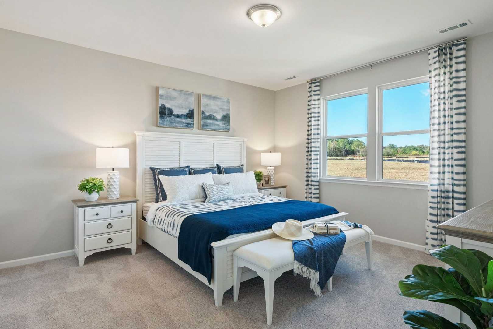 Spacious master bedroom in The Phoenix with white king bed, blue bedding, nightstands, large windows with curtains, and potted plants