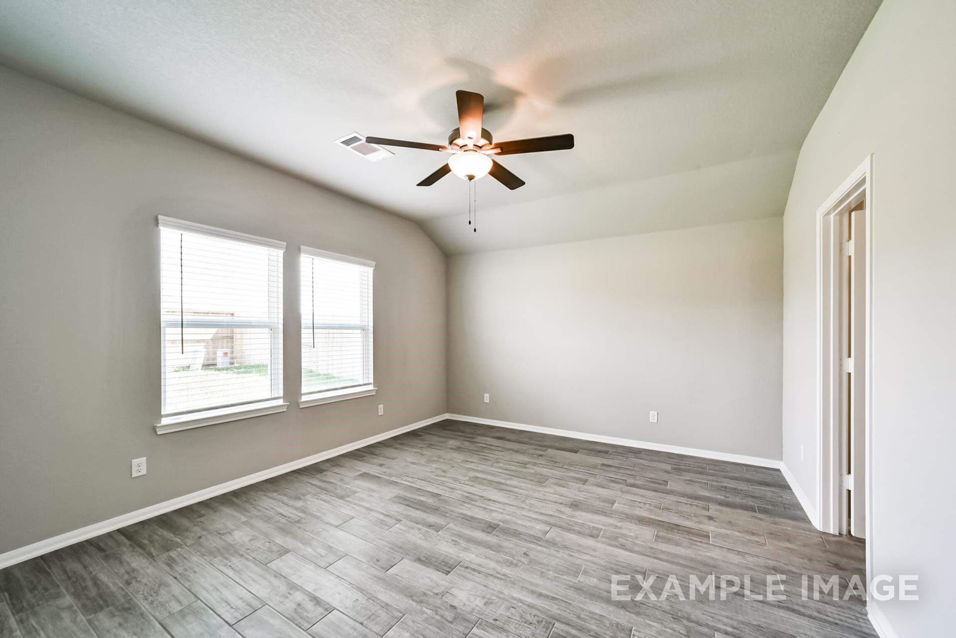 Spacious bedroom with grey wood-look tile flooring and ceiling fan in The Riviera A floor plan at Emberly, Beasley, Texas.