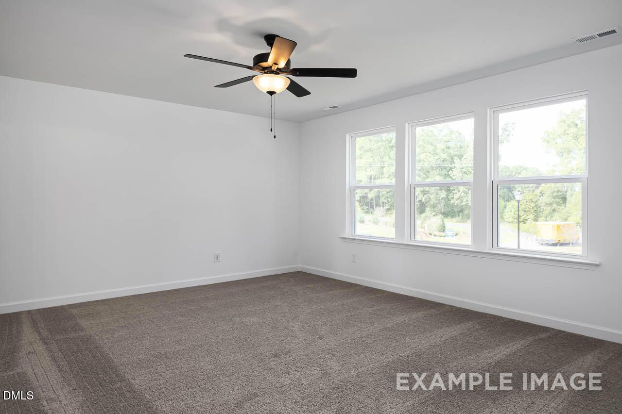 Bright bedroom with large triple windows, ceiling fan, and beige carpet in The Willow D by Davidson Homes, Zebulon, NC
