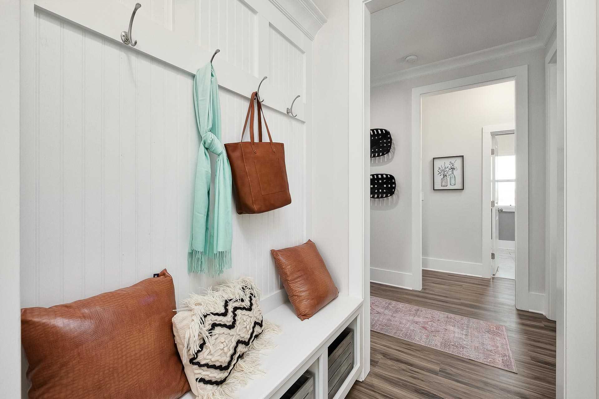 White shiplap mudroom in Newby Chapel Athens AL with hooks, turquoise scarf, tan handbag, cushioned bench, pillows, and hardwood hallway
