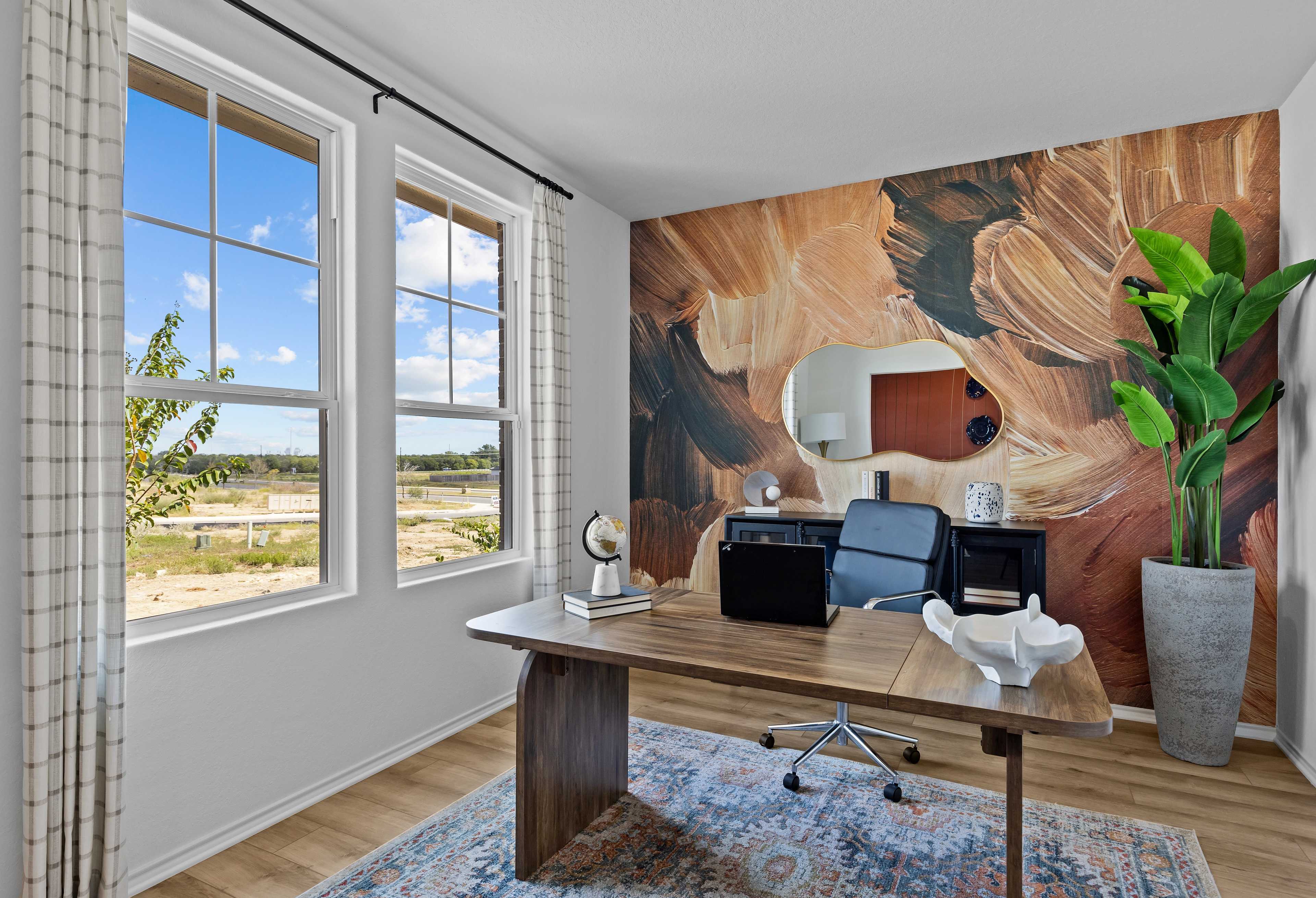 Spacious home office in The Douglas B featuring wooden desk, black chair, abstract earthy mural, potted plant, and large windows overlooking Texas landscape