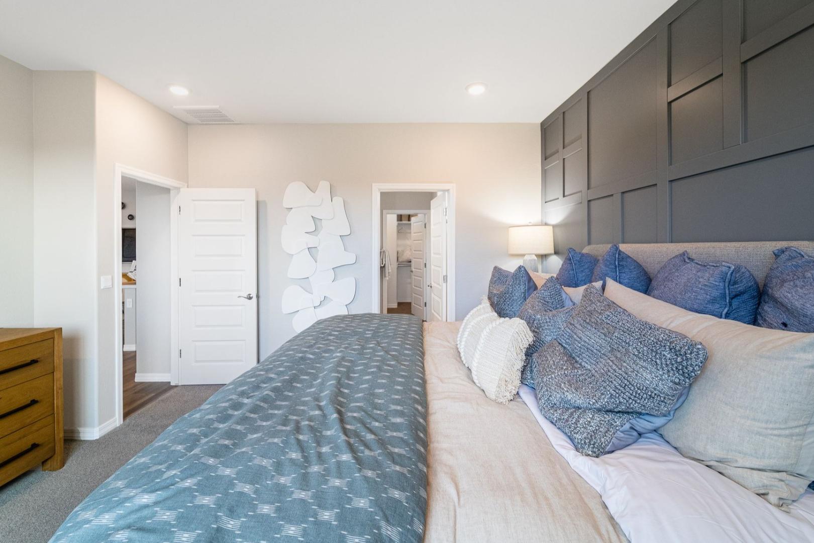 Spacious master bedroom in The Newport C with king bed, blue duvet, plush pillows, wooden dresser, gray accent wall, and ensuite doors
