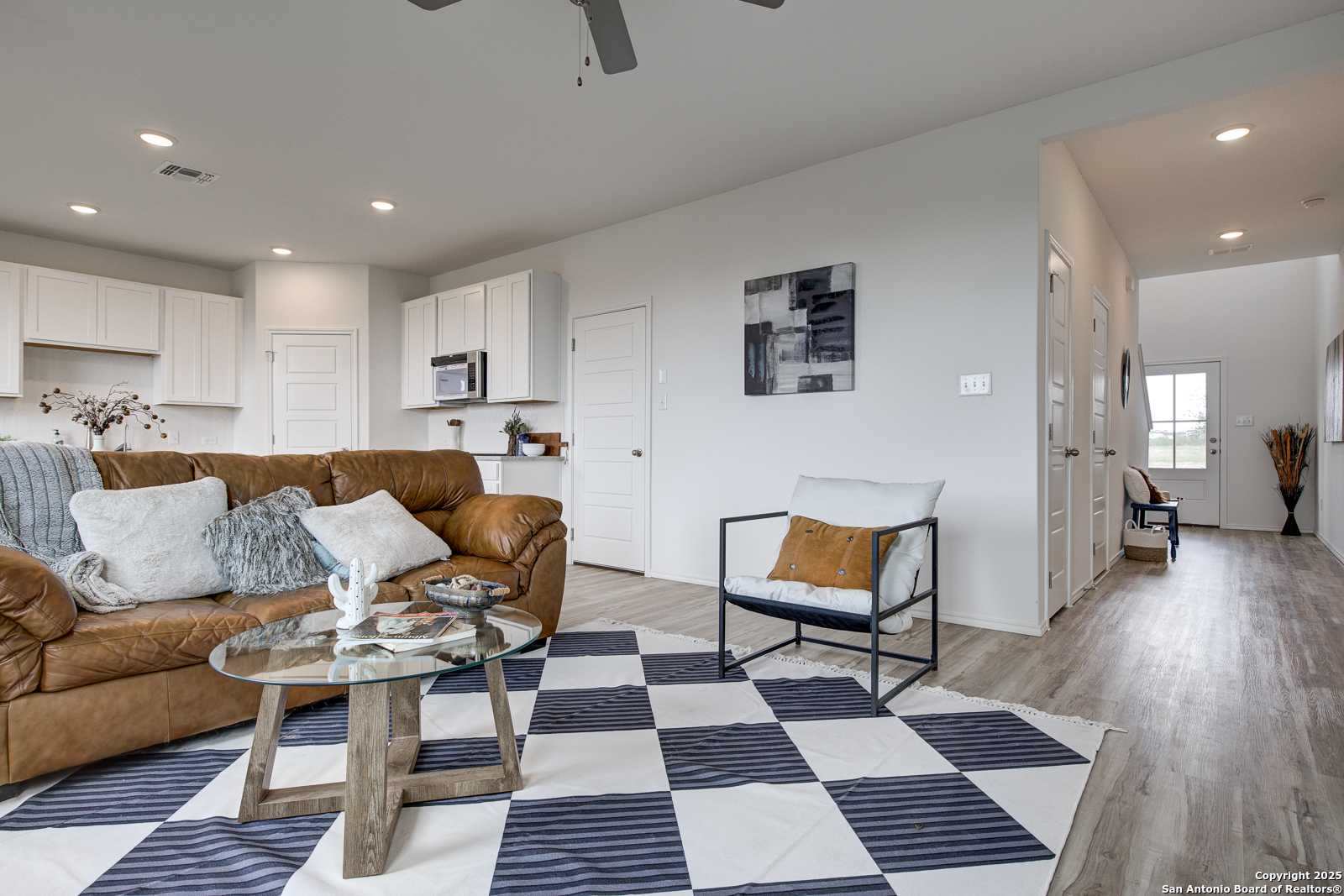 Open-concept living room with leather sofa, glass coffee table, and white kitchen cabinets in Davidson Homes The Trinity A, San Antonio, Texas