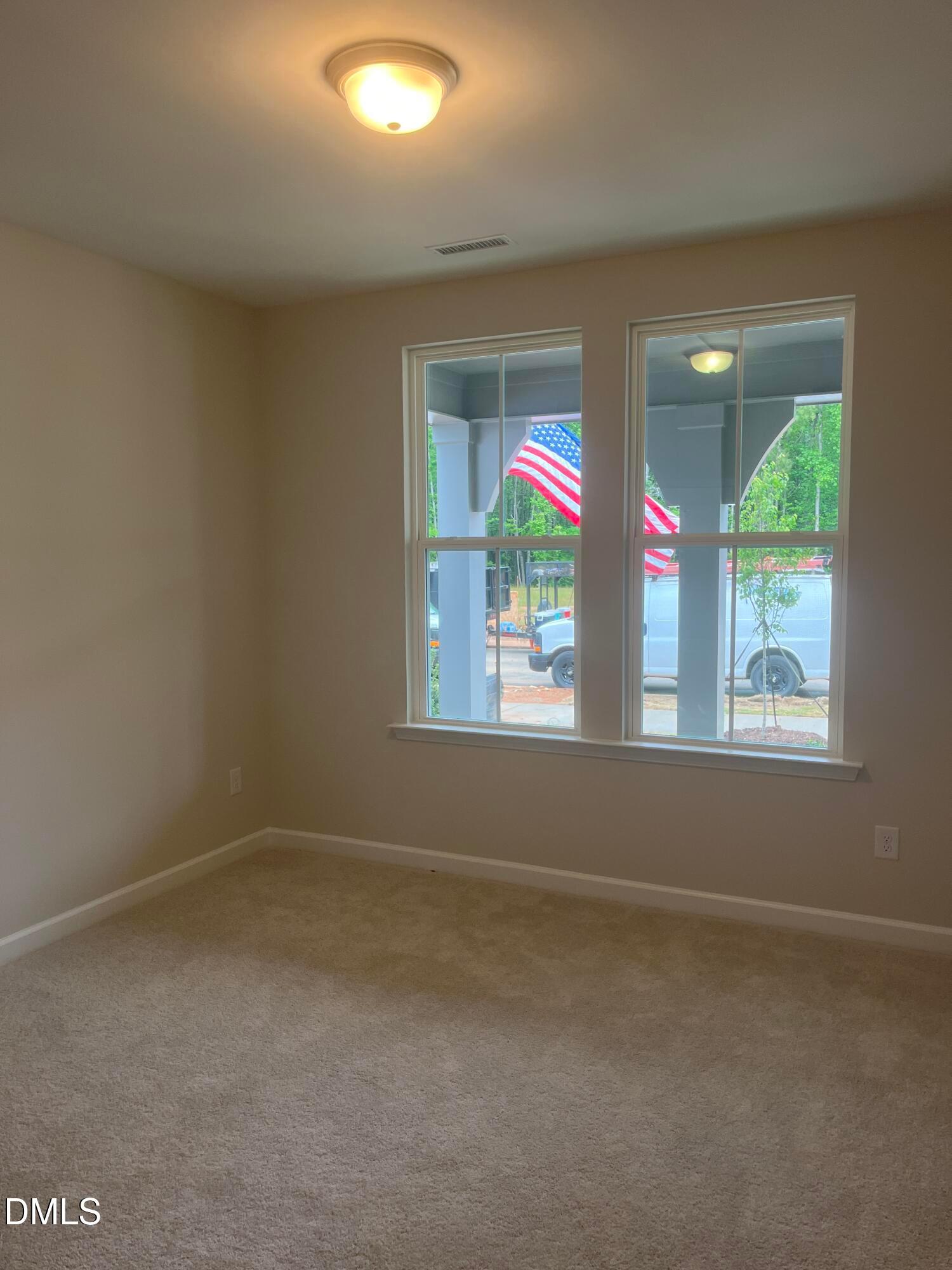 Empty bedroom with beige walls, carpeted floor, and large windows overlooking porch with American flag in Davidson Homes The Ashport G, Wake Forest, NC