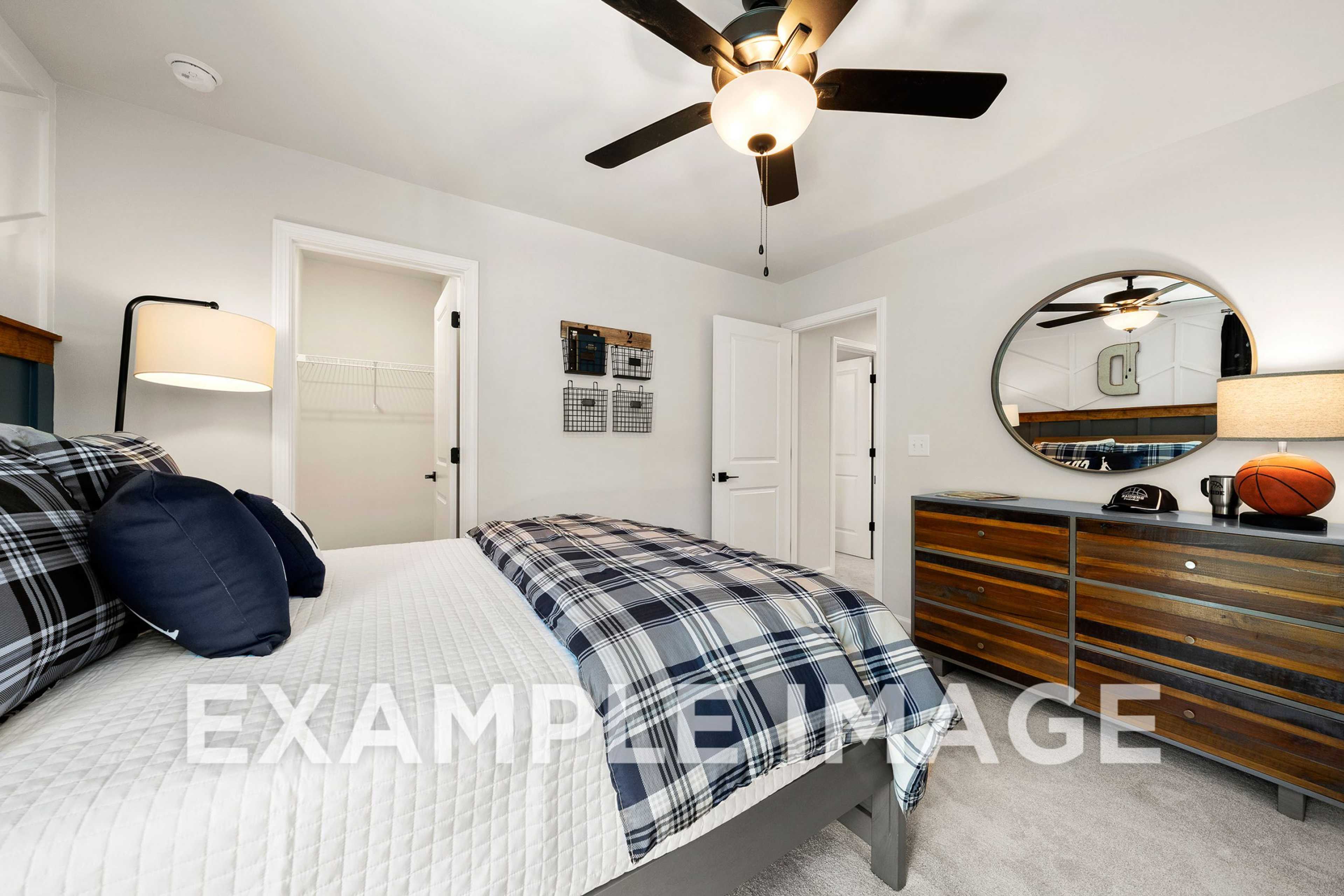 Spacious upper-floor bedroom in The Hickory A by Davidson Homes, featuring plaid bedding, wooden dresser, round mirror, and basketball decor
