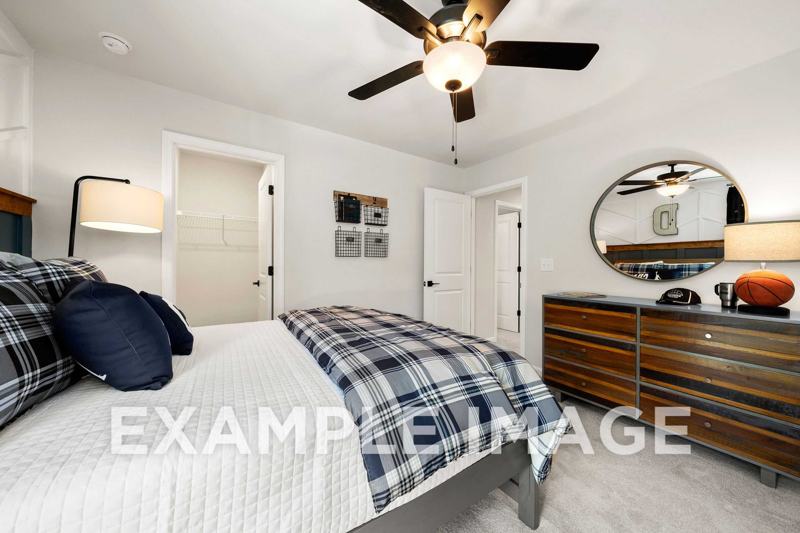 Spacious upper-floor bedroom in The Hickory A by Davidson Homes, featuring plaid bedding, wooden dresser, round mirror, and basketball decor