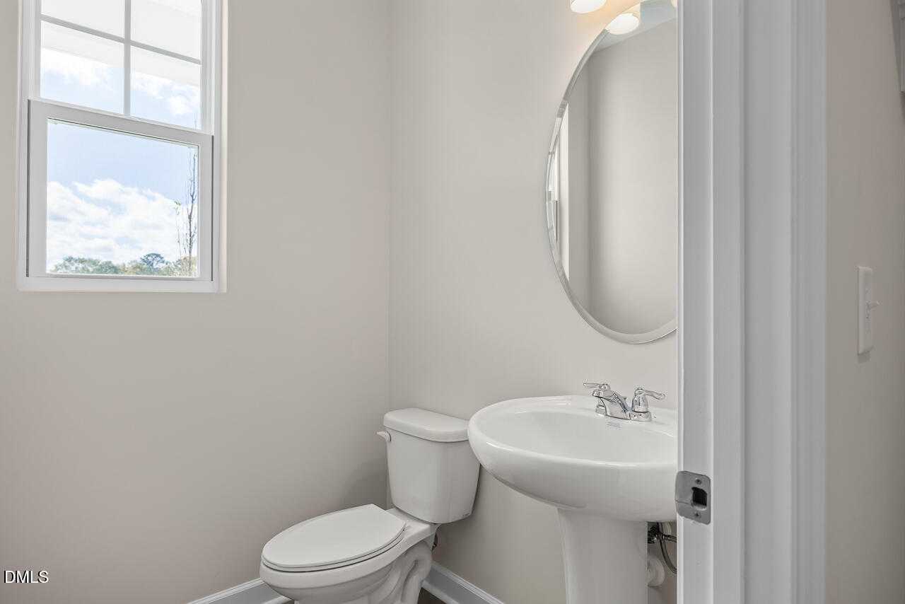 Bright half bath with white sink, toilet, round mirror, and window in Davidson Homes The Graham, Fuquay-Varina, NC