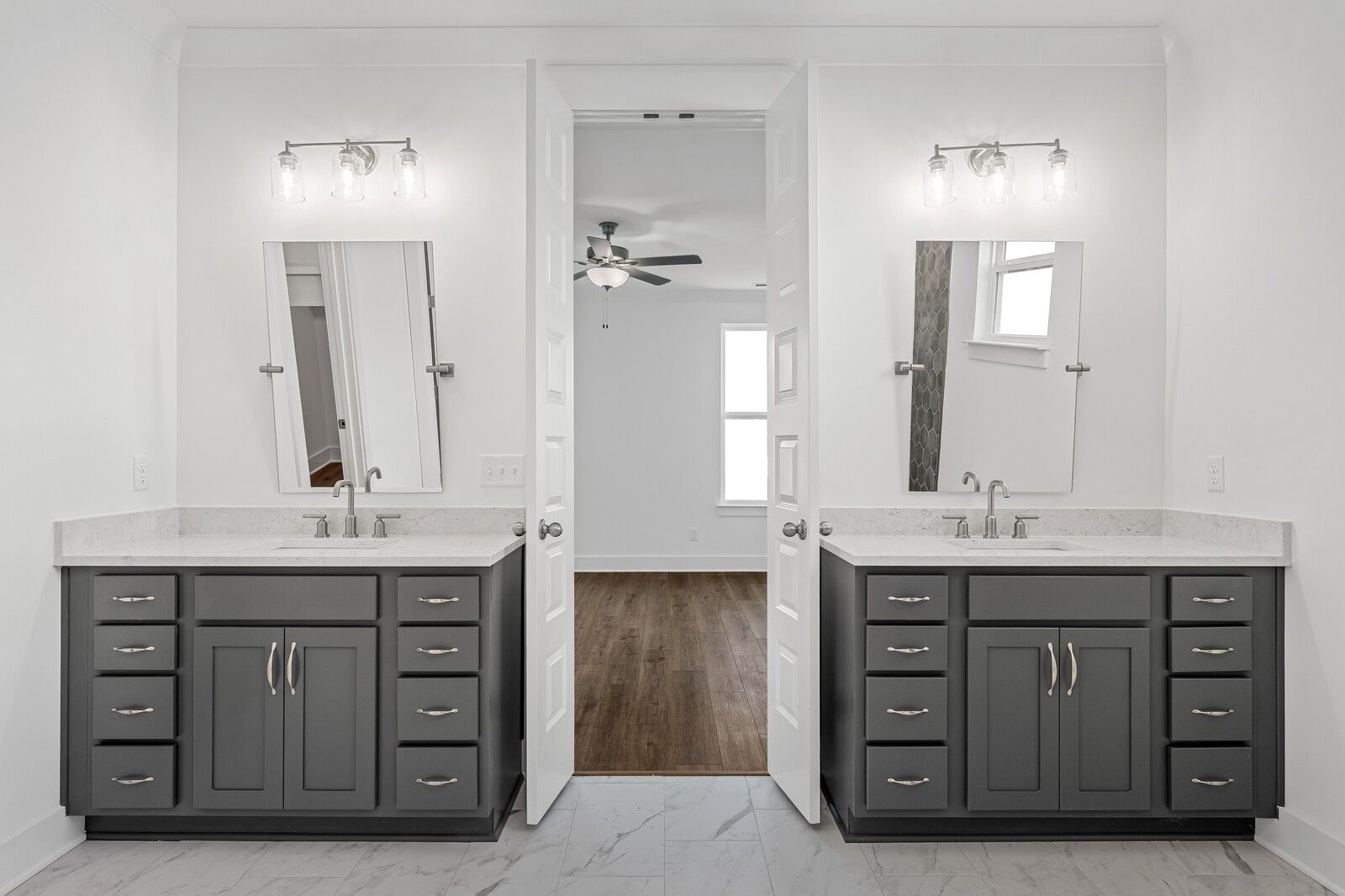 Elegant master bathroom featuring double gray vanities, large mirrors, sconce lights, and open doors to bedroom with ceiling fan in Davidson Homes The Hawkins, Murfreesboro