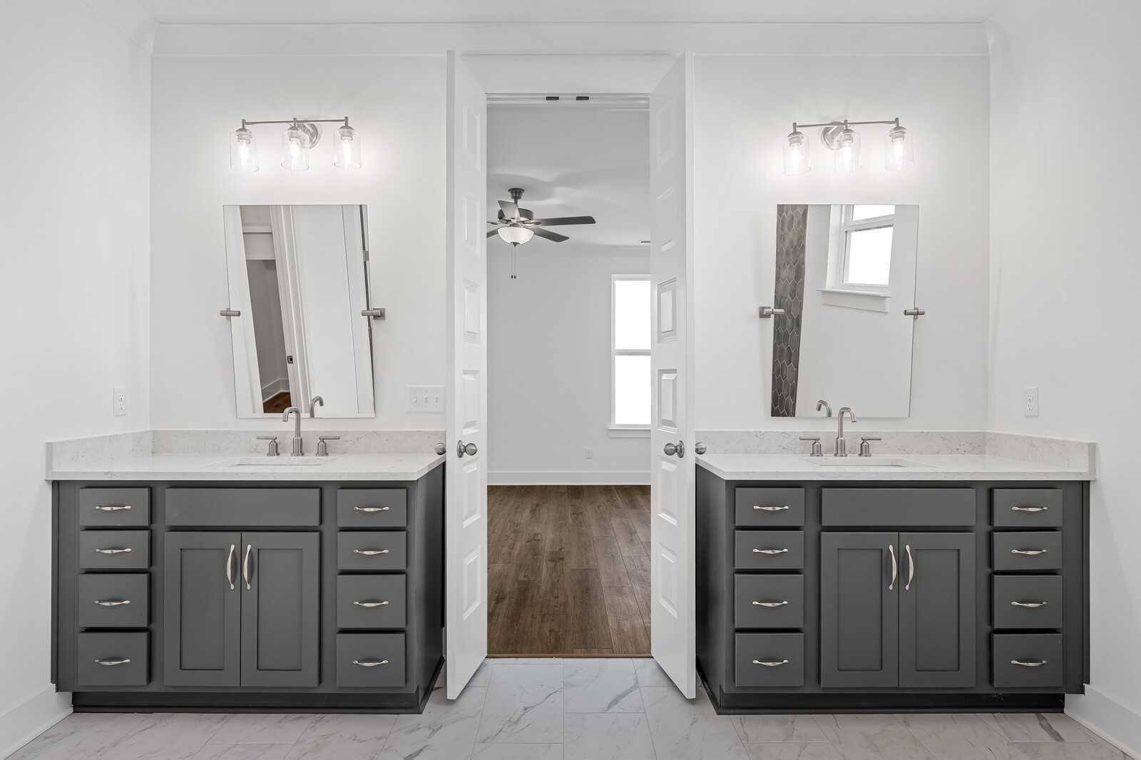 Elegant master bathroom featuring double gray vanities, large mirrors, sconce lights, and open doors to bedroom with ceiling fan in Davidson Homes The Hawkins, Murfreesboro