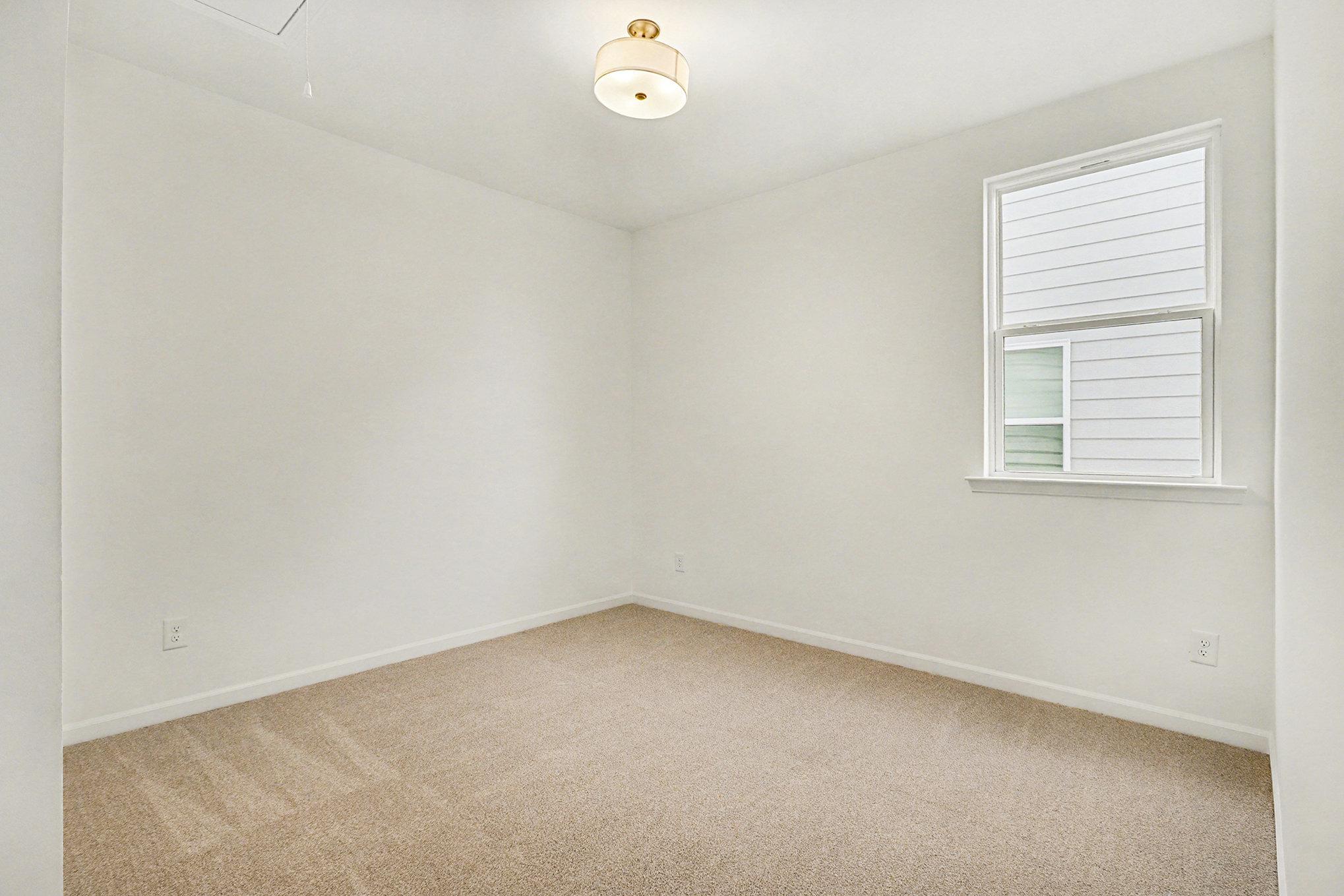 Cozy secondary bedroom with tufted gray headboard bed, rainbow duvet, nightstand lamp, and window in Davidson Homes The Daphne B, Loganville, GA