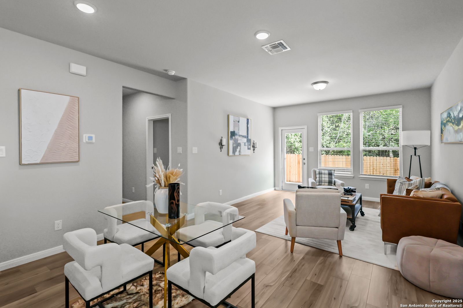 Modern open dining-living space with glass table, white chairs, beige sofa, large windows, hardwood floors in Davidson Homes The Gillian C, San Antonio