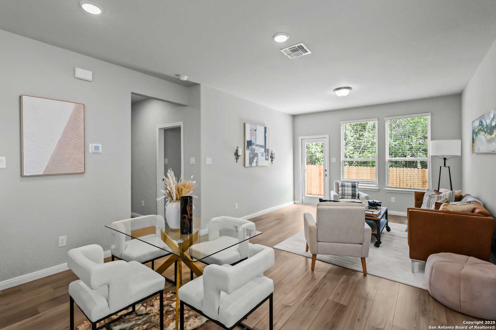 Modern open dining-living space with glass table, white chairs, beige sofa, large windows, hardwood floors in Davidson Homes The Gillian C, San Antonio