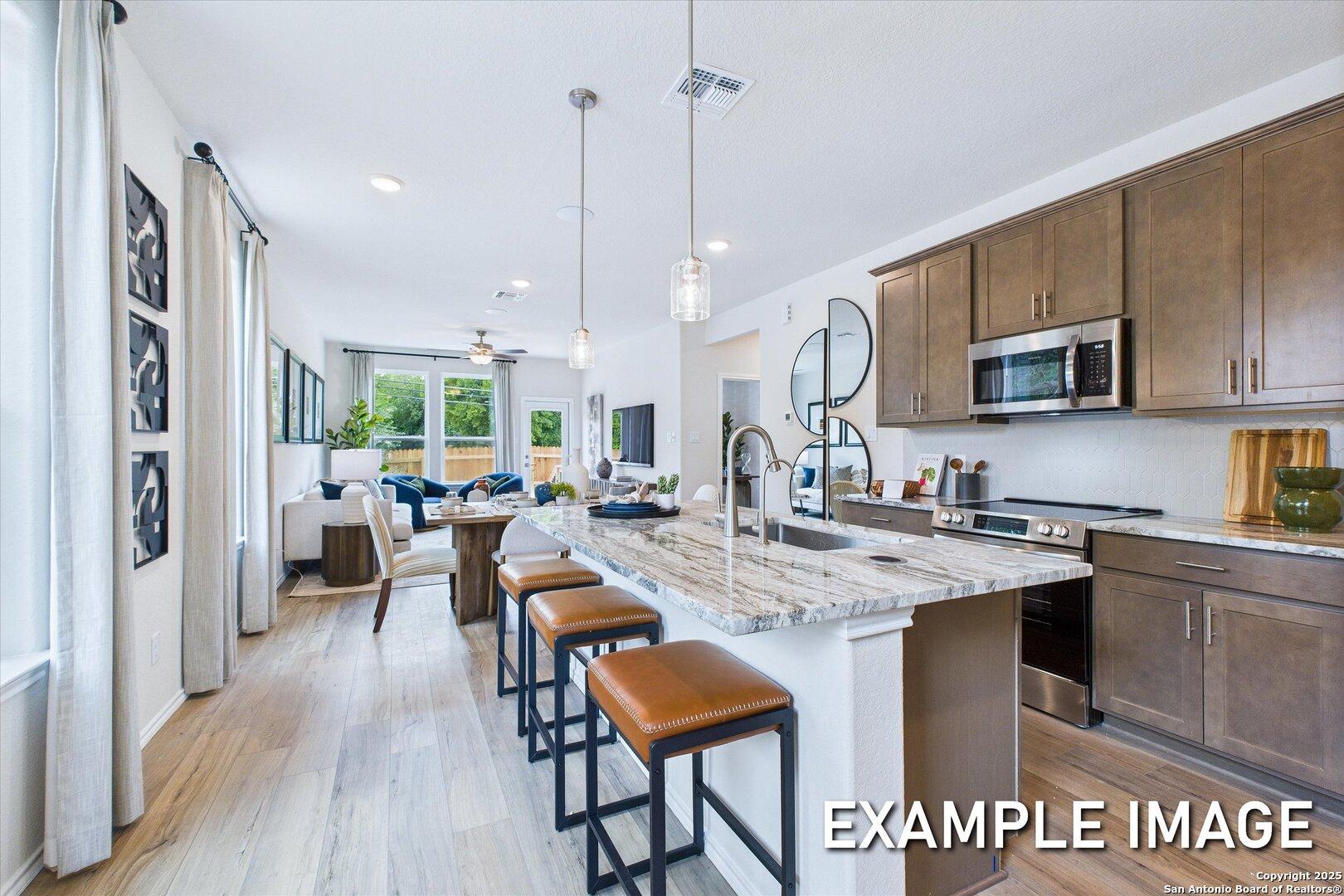 Modern open-concept kitchen with quartz island, brown cabinets, stainless appliances, and adjacent living area in The Gillian C home, San Antonio