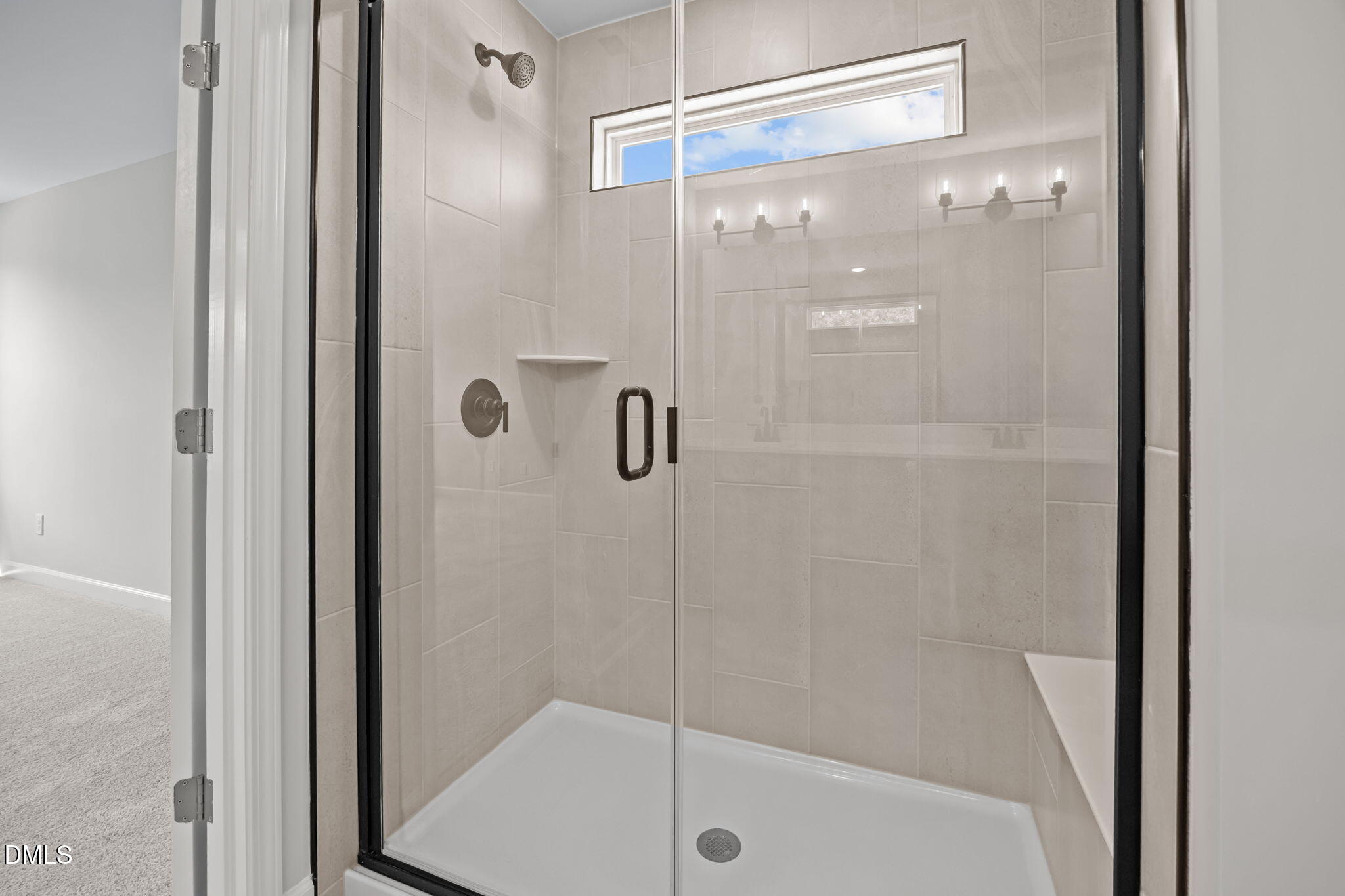 Modern frameless glass walk-in shower with light tile walls, rainfall head, bench seat, and transom window in The Willow D master bath, Wendell NC