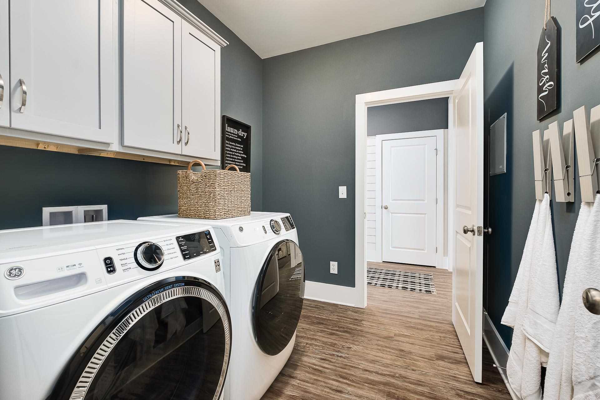 Spacious laundry room in The Everett C with white washer dryer, gray walls, upper cabinets, and hanging towels