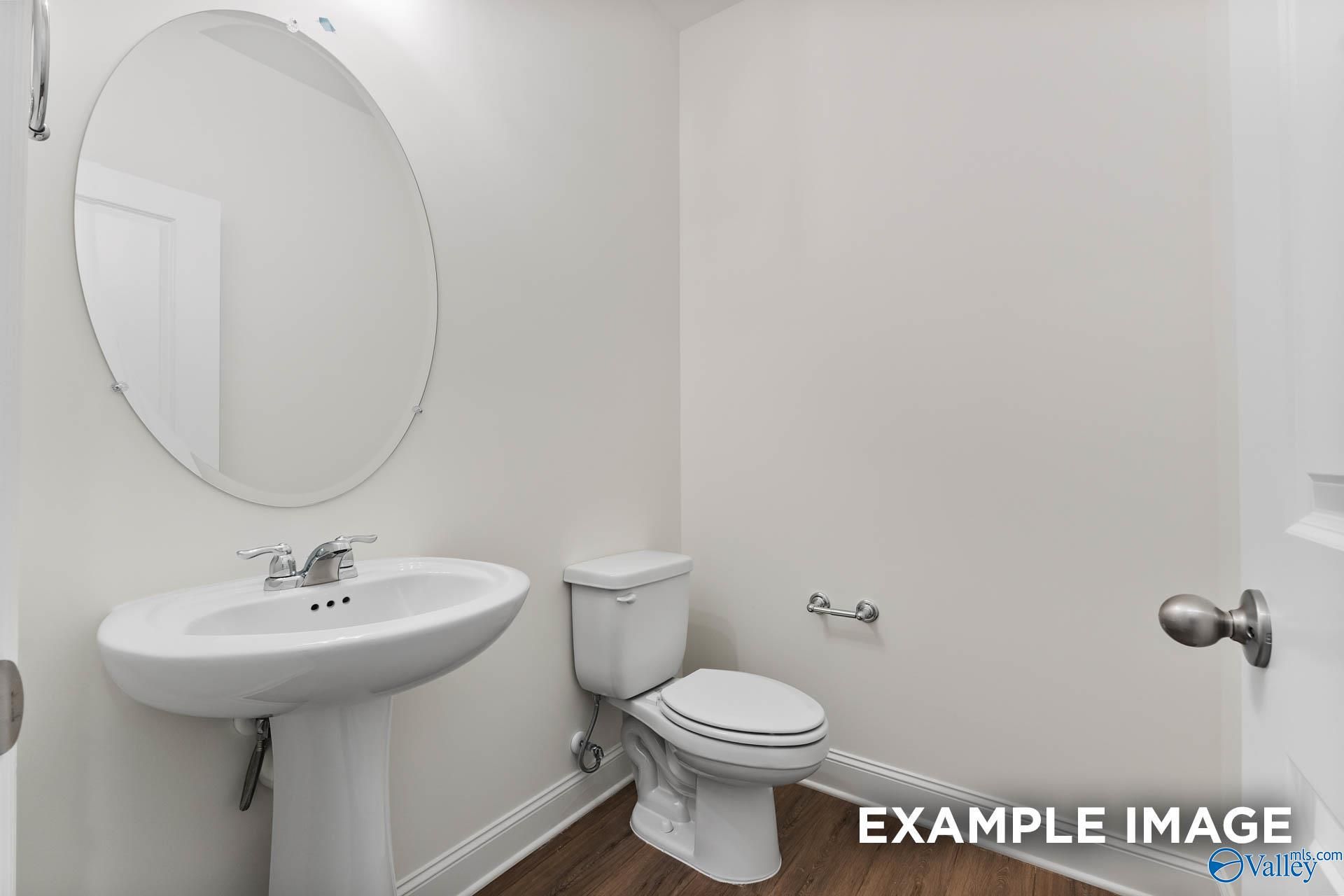 Elegant white powder room with round mirror, pedestal sink, and toilet on hardwood floors in Davidson Homes The Harrison C, Athens, Alabama