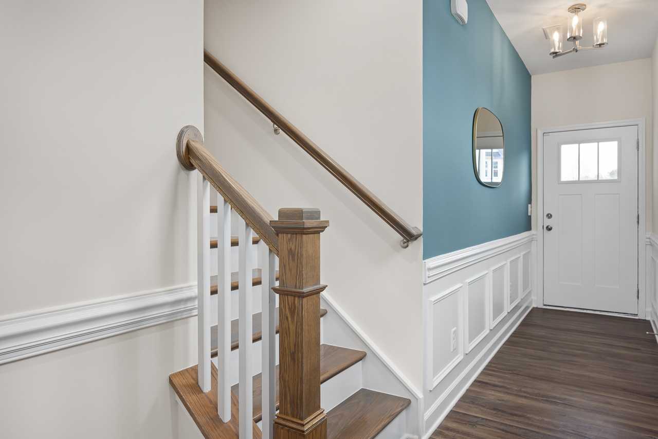 Spacious entryway featuring wooden staircase, teal accent wall, round mirror, and hardwood floors at Gregory Village Townhomes in Lillington NC