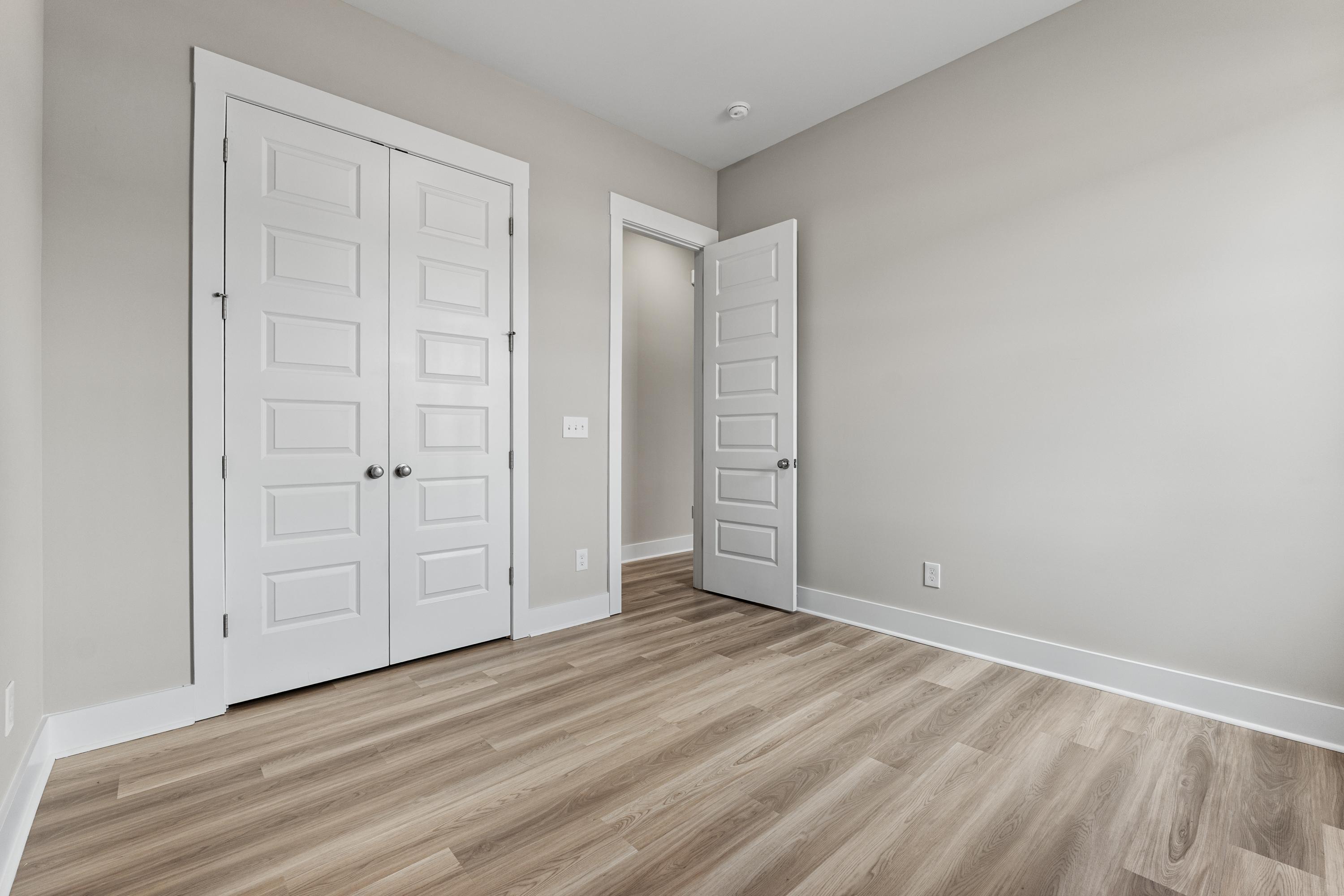 Spacious bedroom in The Oxford C with light gray walls, white double closet doors, open bath doorway, and light oak hardwood floors