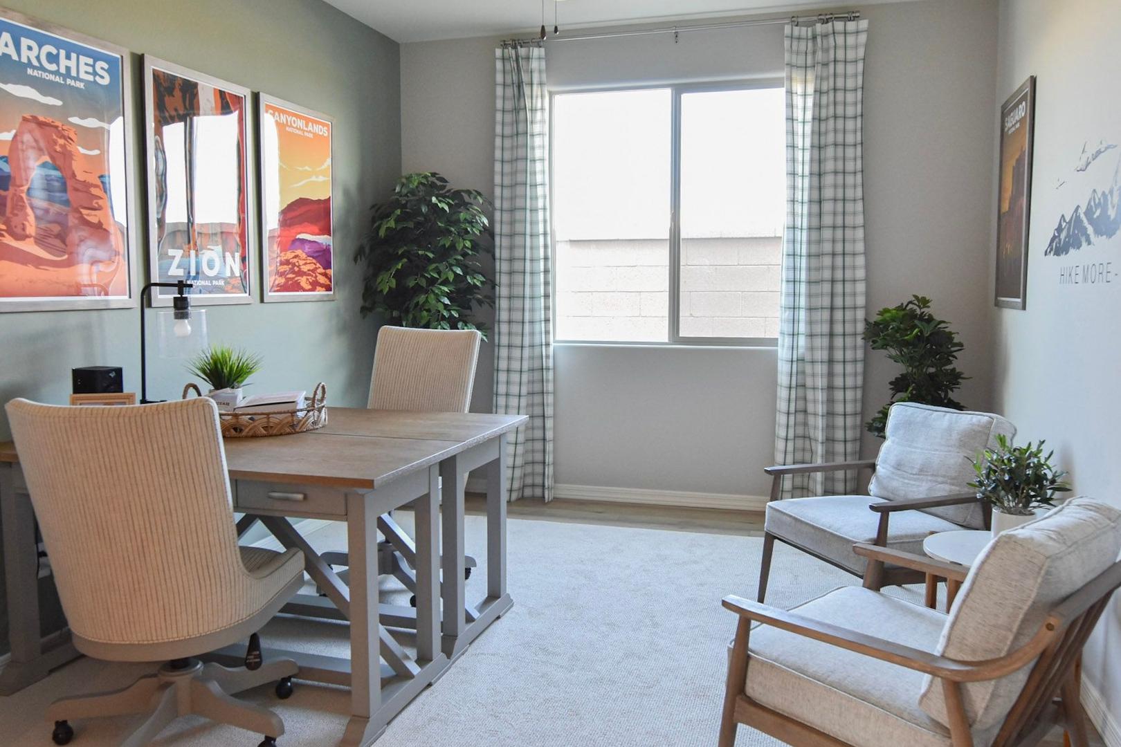 Cozy home office in The Harmony B featuring desert landscape wall art, wooden desk with task chairs, potted plants, and large window