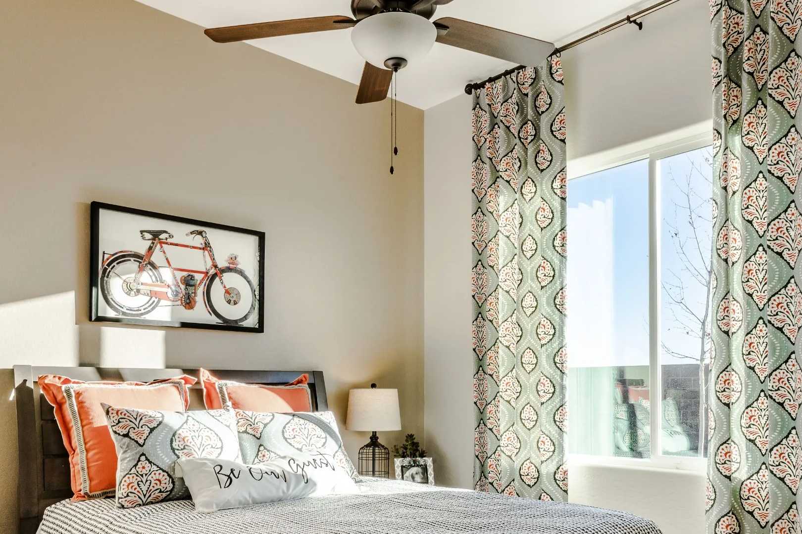 Cozy bedroom interior in Westwood Prescott AZ by Davidson Homes with queen bed orange pillows ceiling fan teal curtains and bicycle wall art
