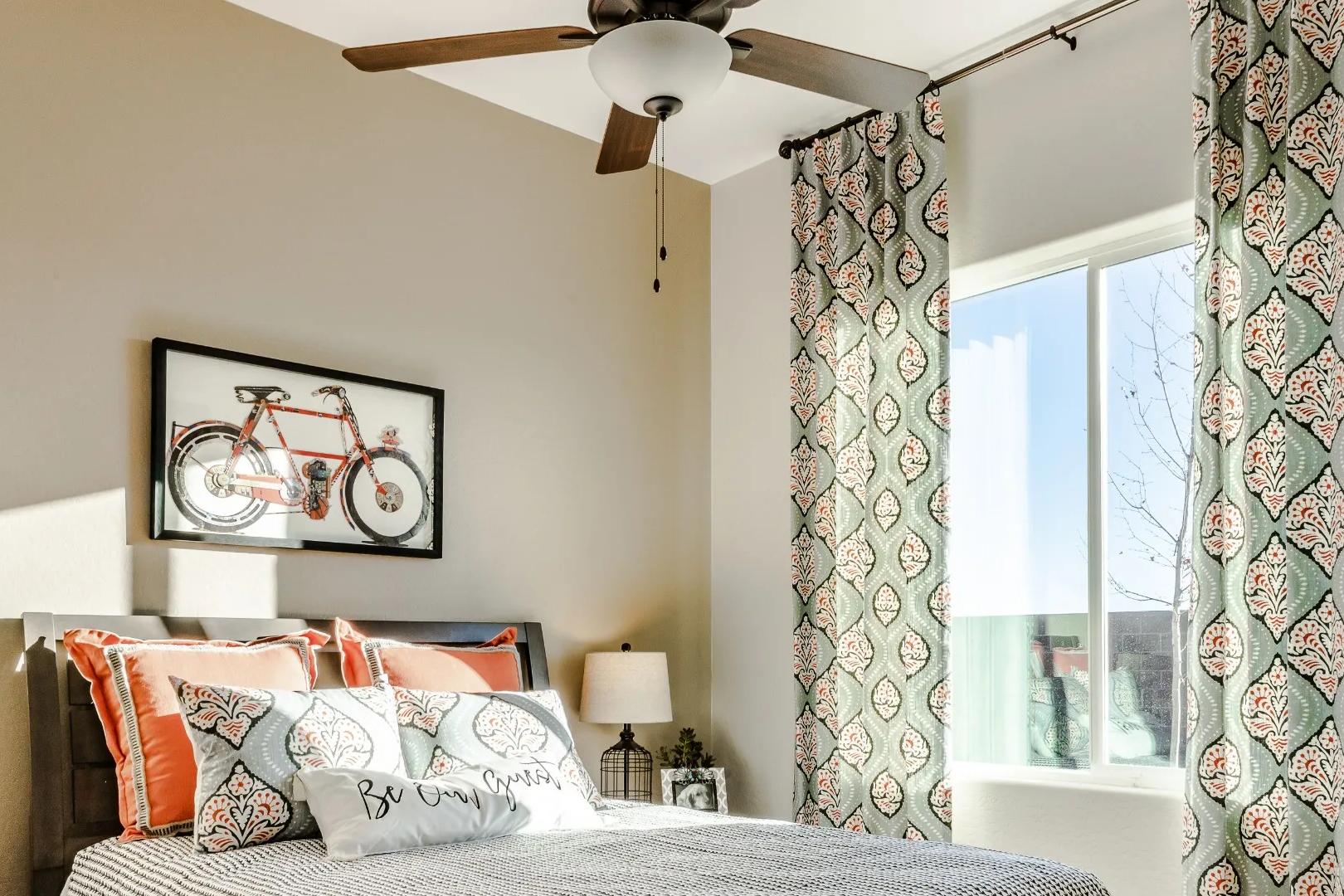 Cozy bedroom interior in Westwood Prescott AZ by Davidson Homes with queen bed orange pillows ceiling fan teal curtains and bicycle wall art