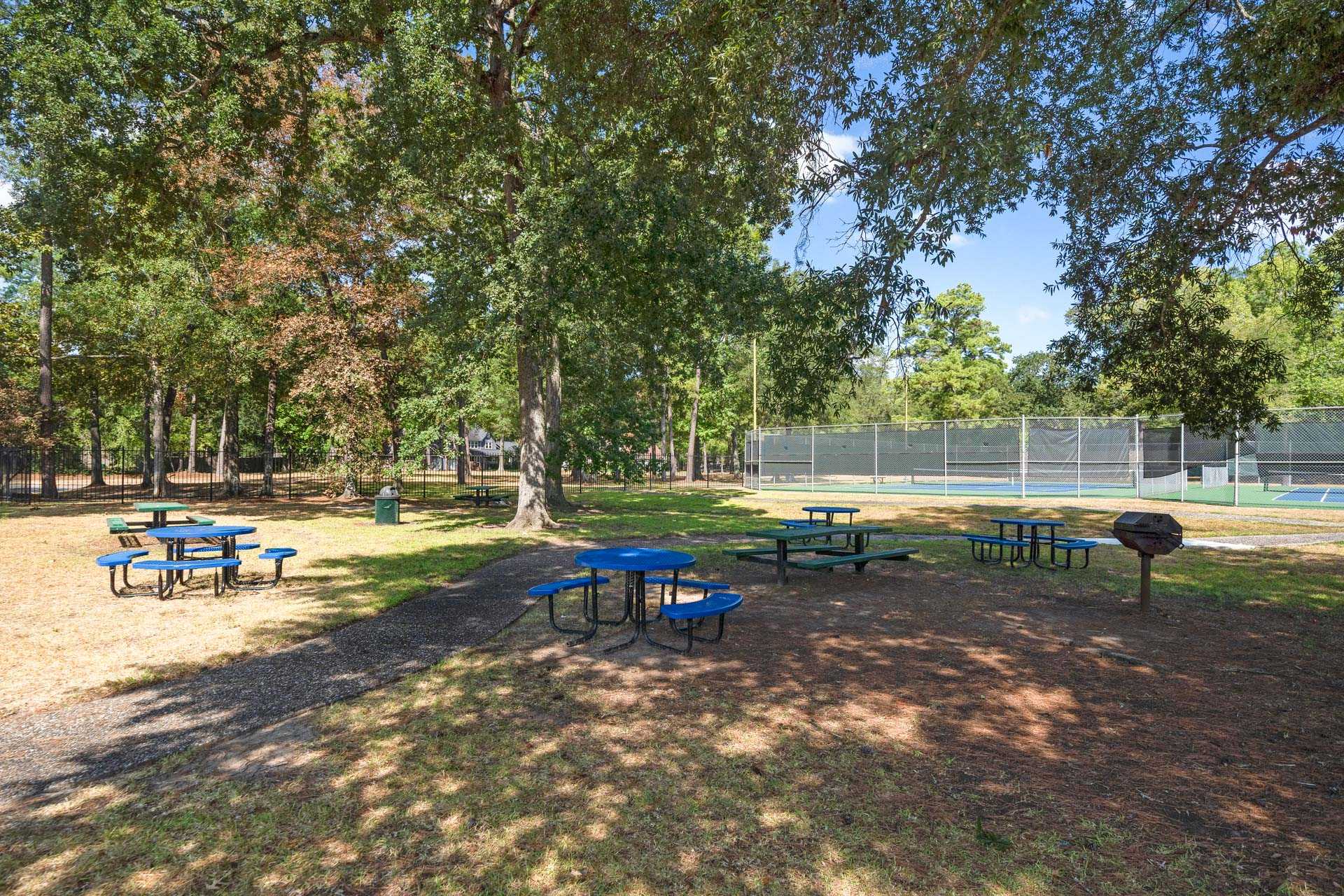 Tree-lined community park at Enclave at Newport in Crosby Texas with blue picnic tables, benches, grill and fenced tennis courts