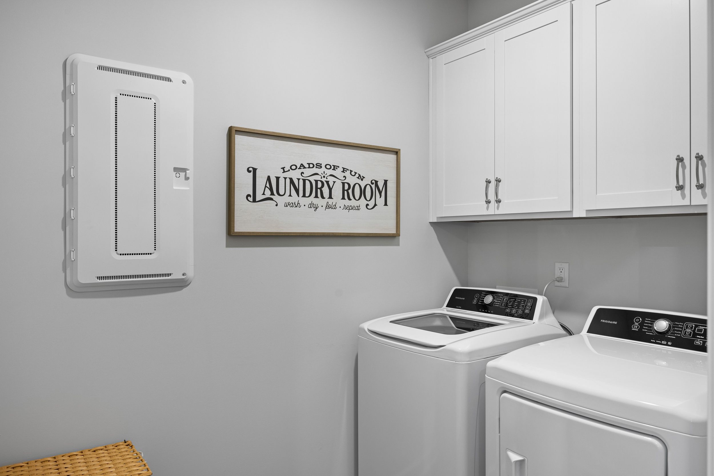 Modern laundry room in The Magnolia B featuring white washer-dryer set, upper cabinets, and "Loads of Fun" sign