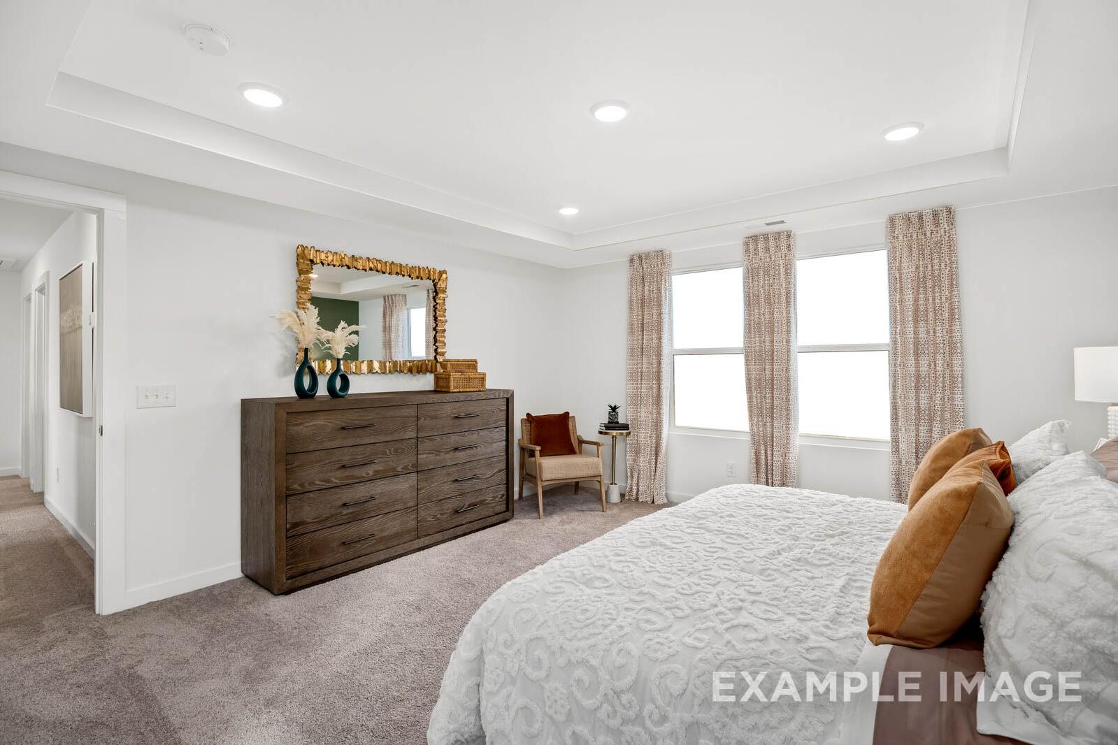 Spacious master bedroom in The Gordon C featuring king bed with plush white bedding, wooden dresser, gold mirror, and large curtained windows