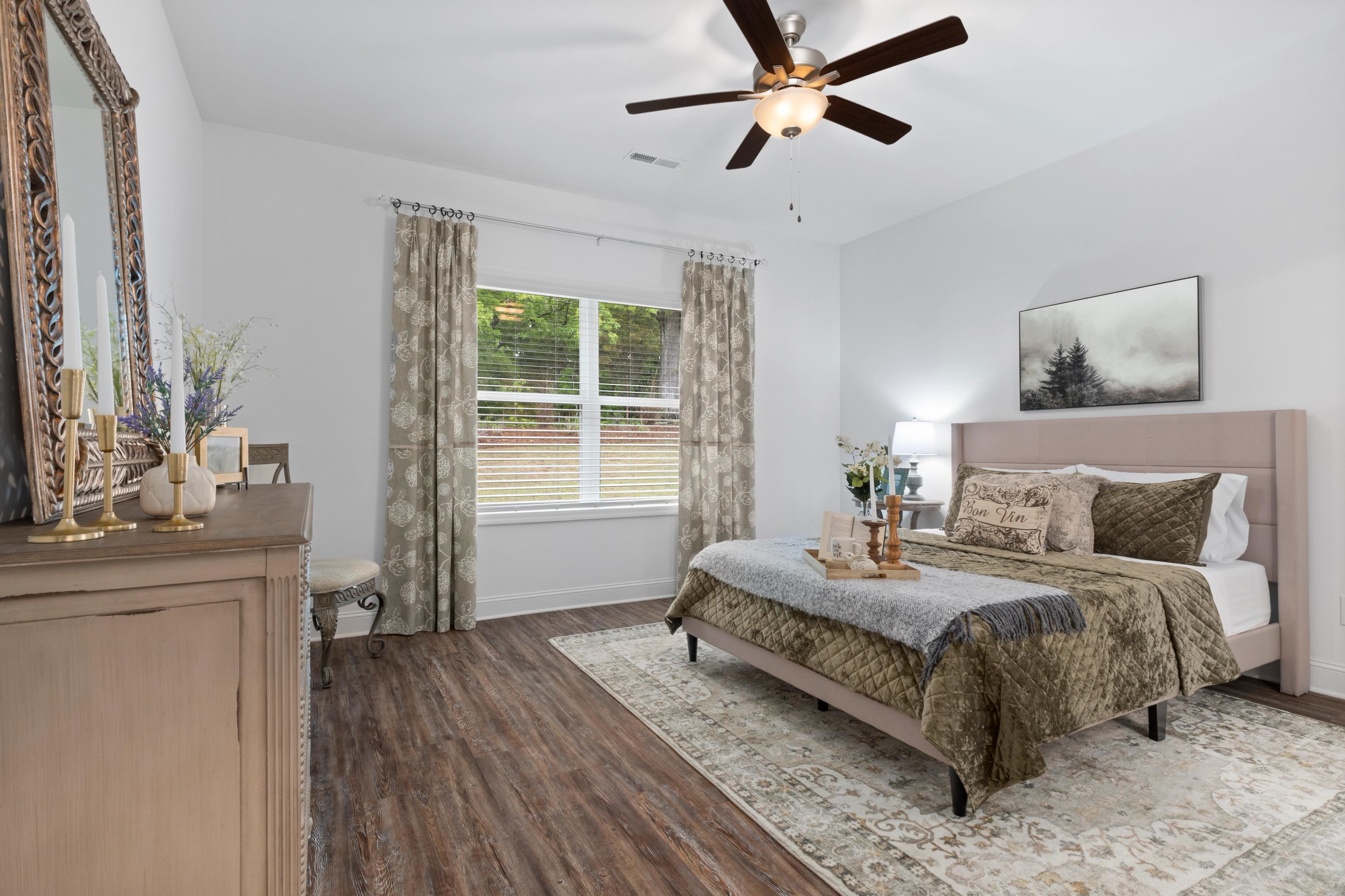 Serene master bedroom in The Cumberland B featuring plush king bed with neutral bedding, wooden dresser, ornate mirror, ceiling fan, and large window with curtains