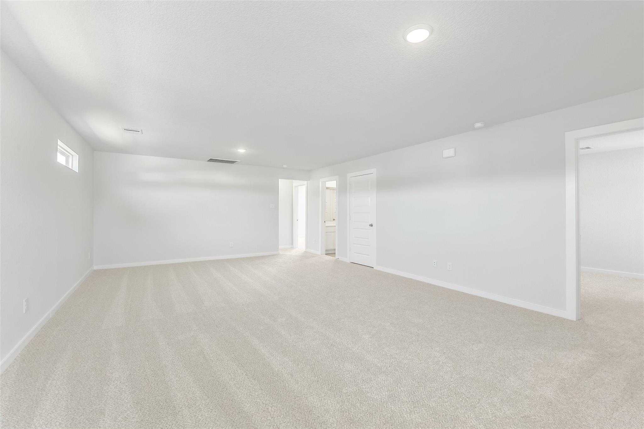 Spacious empty bedroom with beige carpet, white walls, recessed lights, and bathroom doorway in The San Marcos E by Davidson Homes, Cleveland, Texas