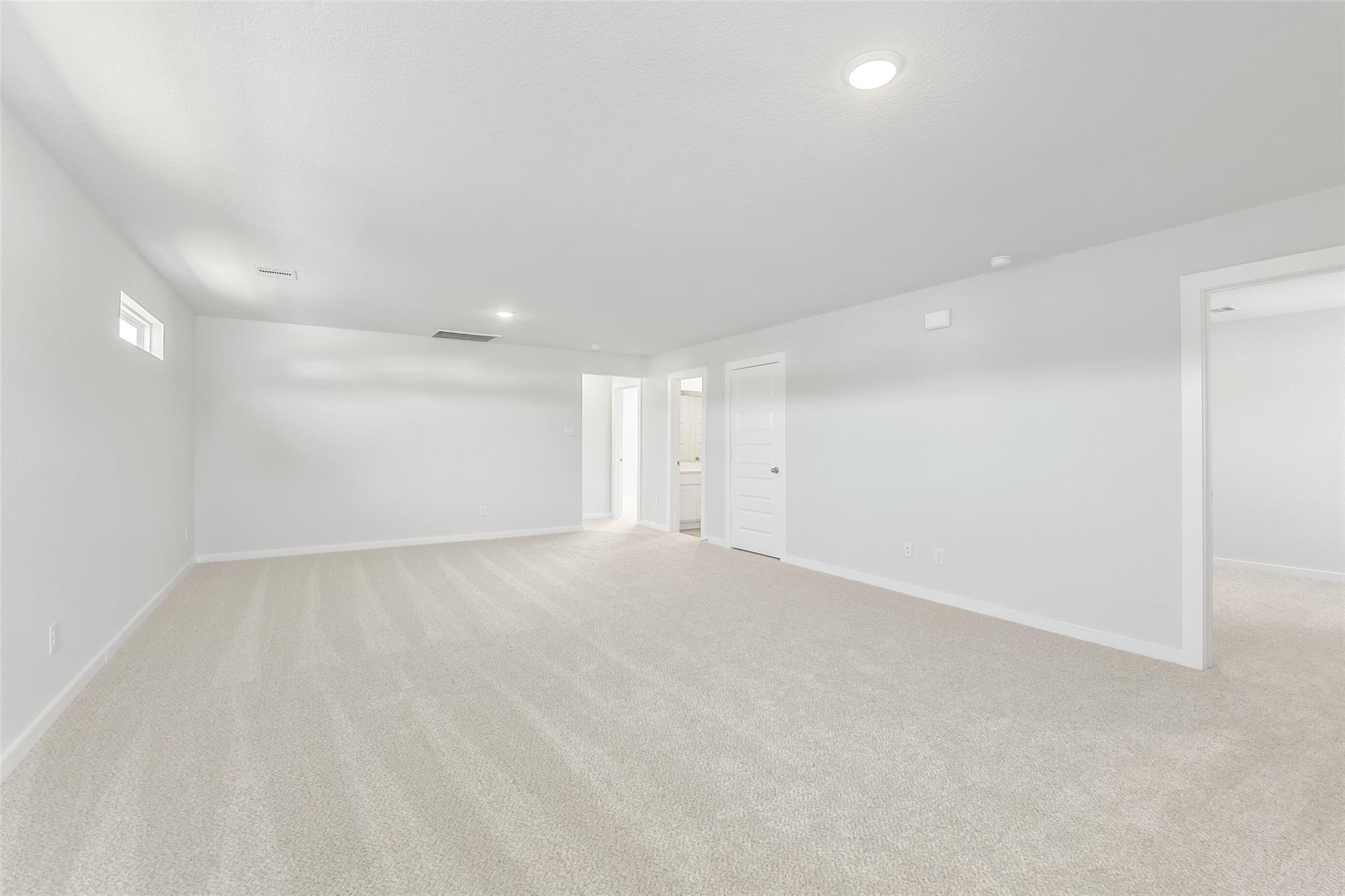 Spacious empty bedroom with beige carpet, white walls, recessed lights, and bathroom doorway in The San Marcos E by Davidson Homes, Cleveland, Texas