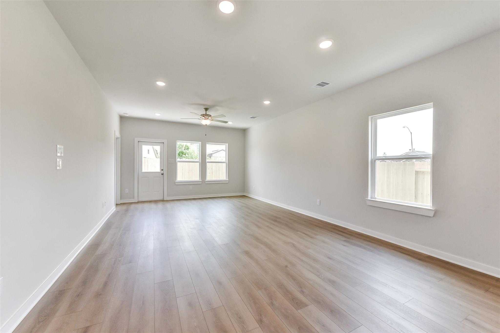 Spacious living room with light hardwood floors, ceiling fan, recessed lights, and large windows to fenced backyard in Davidson Homes The Frio F, Dayton, Texas