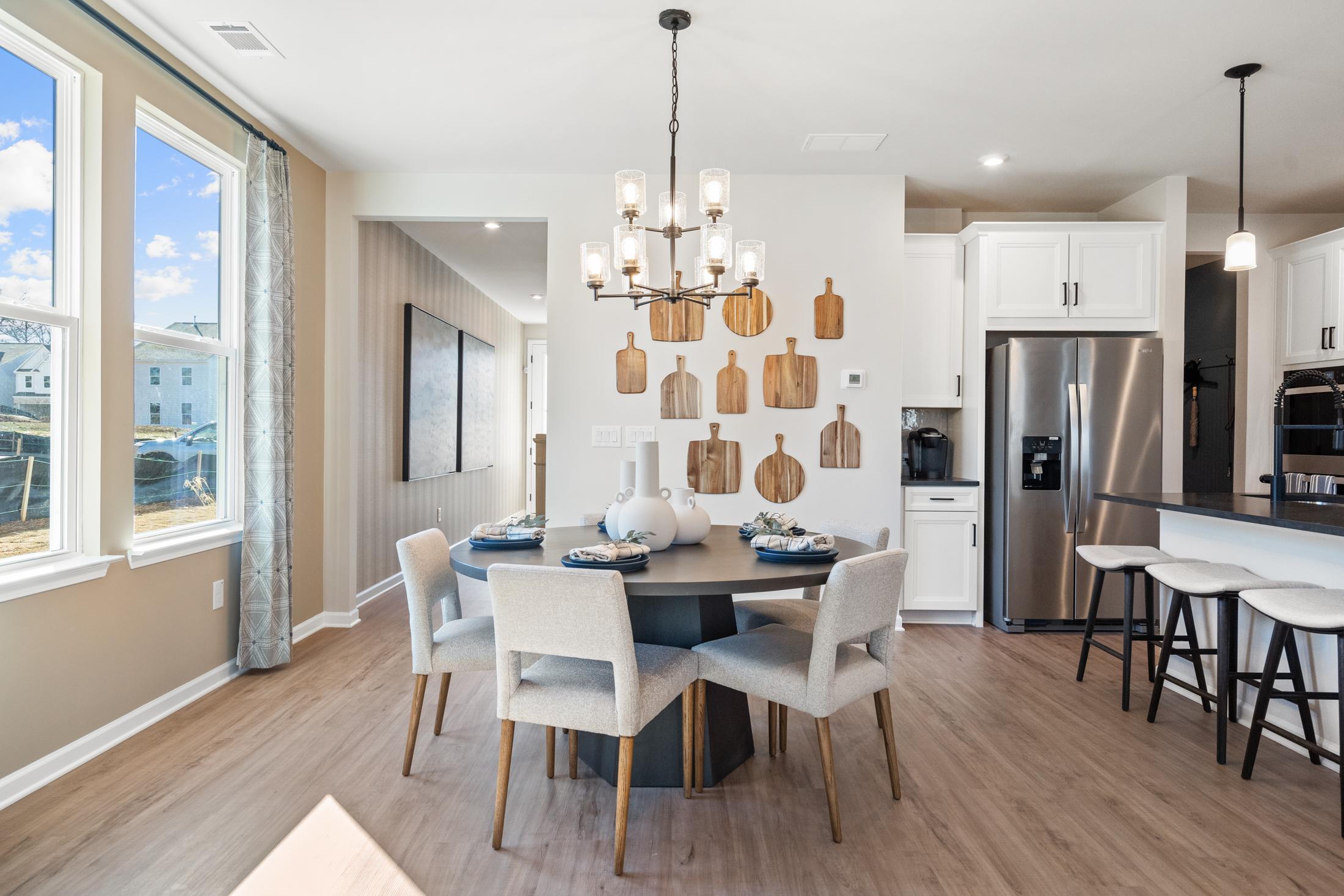 Spacious dining area in The Marion A showcasing round wooden table, upholstered chairs, chandelier, wooden wall accents, and adjacent white kitchen