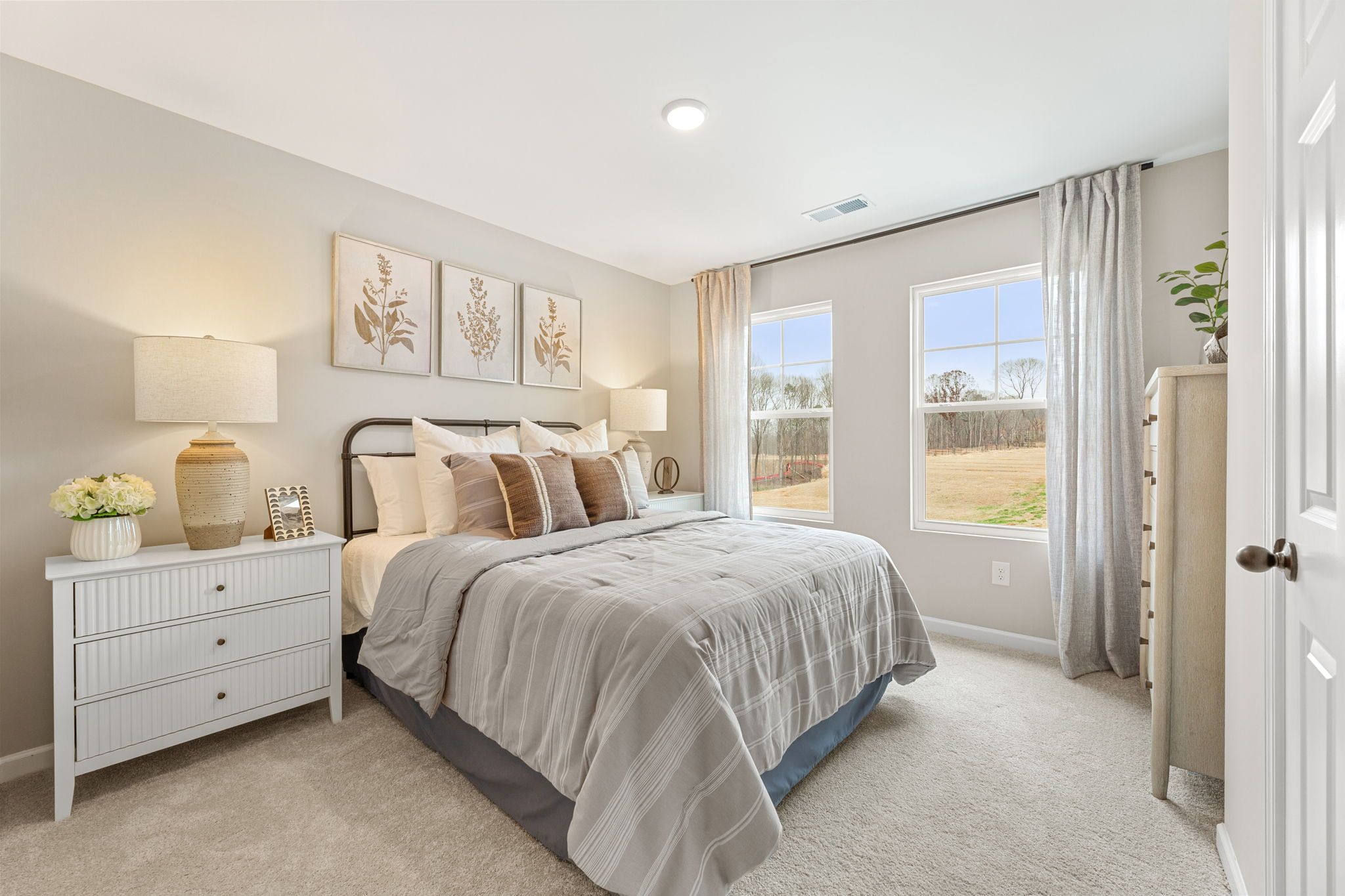 Spacious master bedroom in Hemingway Cumming GA with neutral beige walls, iron bed frame, botanical art, and wooded window views