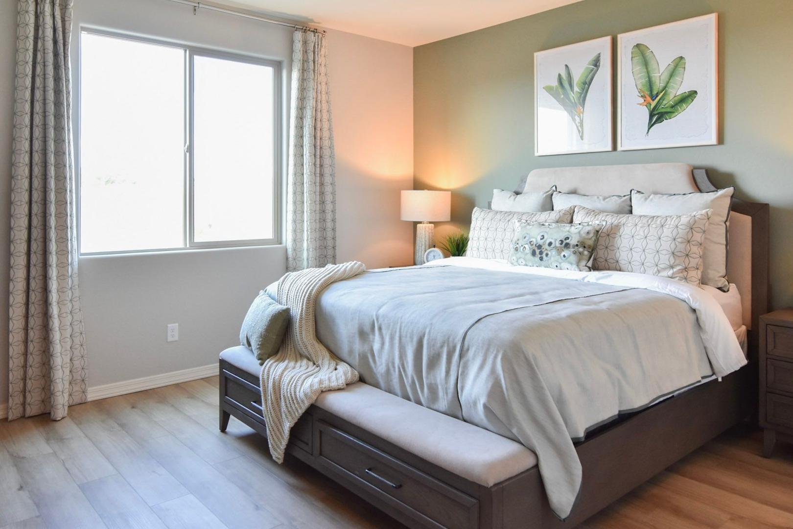 Spacious master bedroom in The Harmony B featuring king bed, sage green wall art, potted plant, and sunlit window