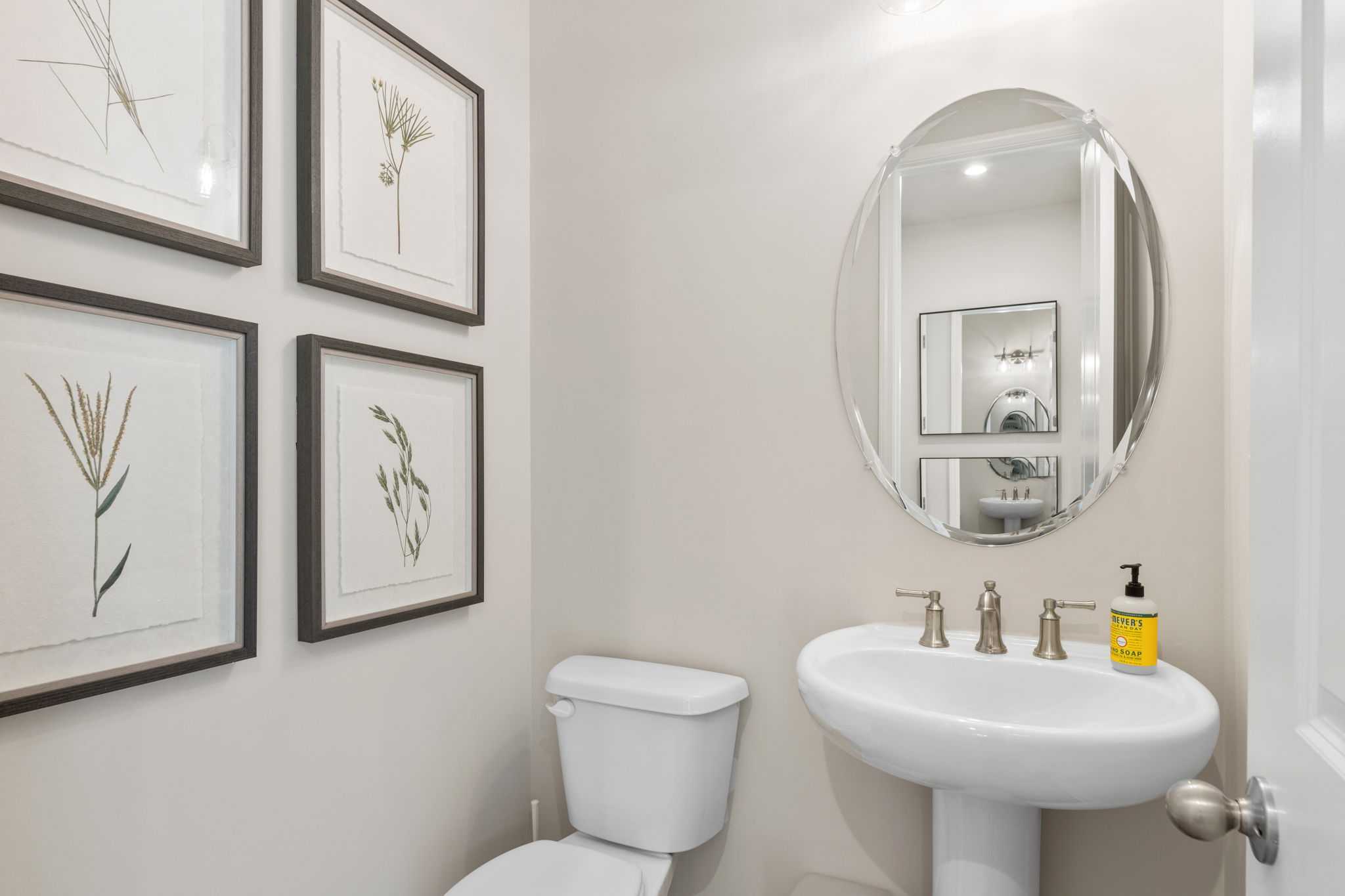 Elegant powder room in Melody Lakeside Estates, Buford GA with pedestal sink, round mirror, and botanical art by Davidson Homes
