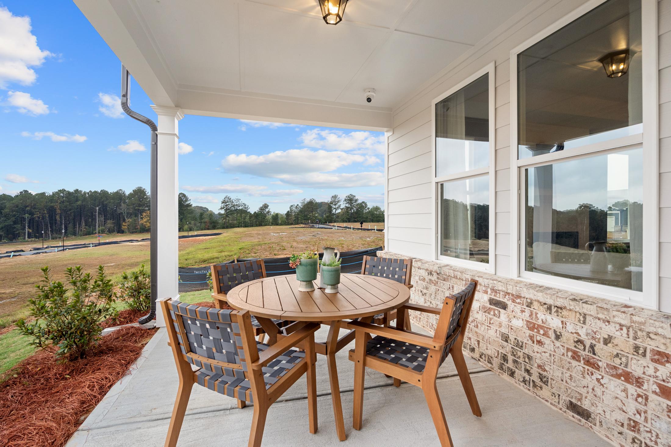 Spacious covered porch of The Edison A home with round teak table, wicker chairs, plants, and scenic golf course view in Loganville, GA