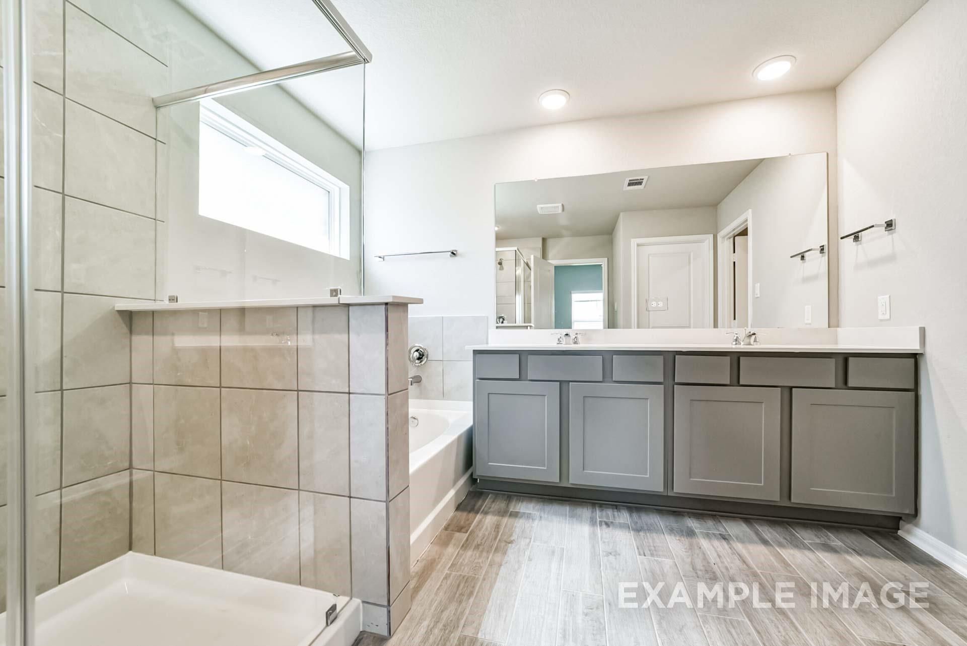 Modern primary bathroom with dual-sink grey vanity, tiled walk-in shower, and soaking tub in The Riviera A at Emberly.