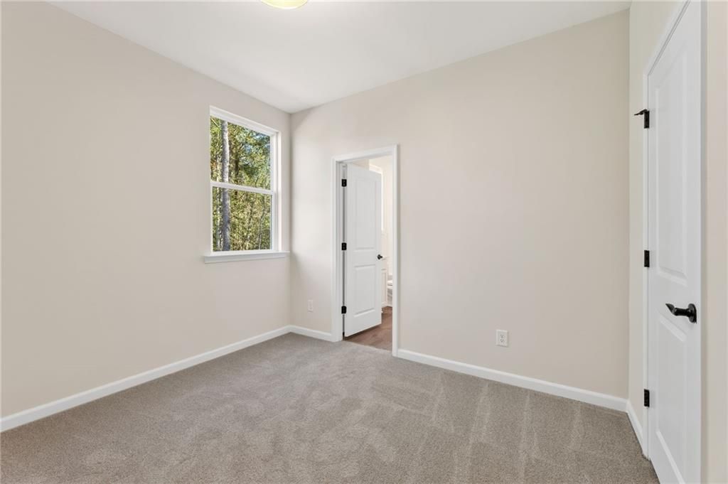 Bright bedroom with beige walls, gray carpet, large window, and en-suite bath door in The Daphne B, Loganville, Georgia