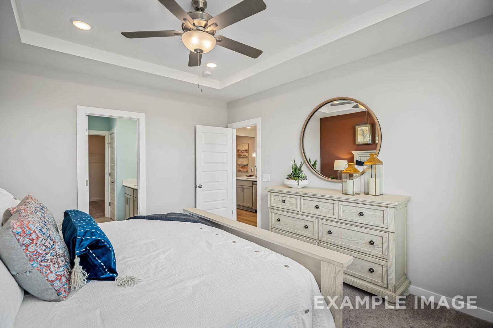 Spacious master bedroom in The Franklin B with white walls, ceiling fan, round mirror on dresser, plush bed, and en-suite bath door