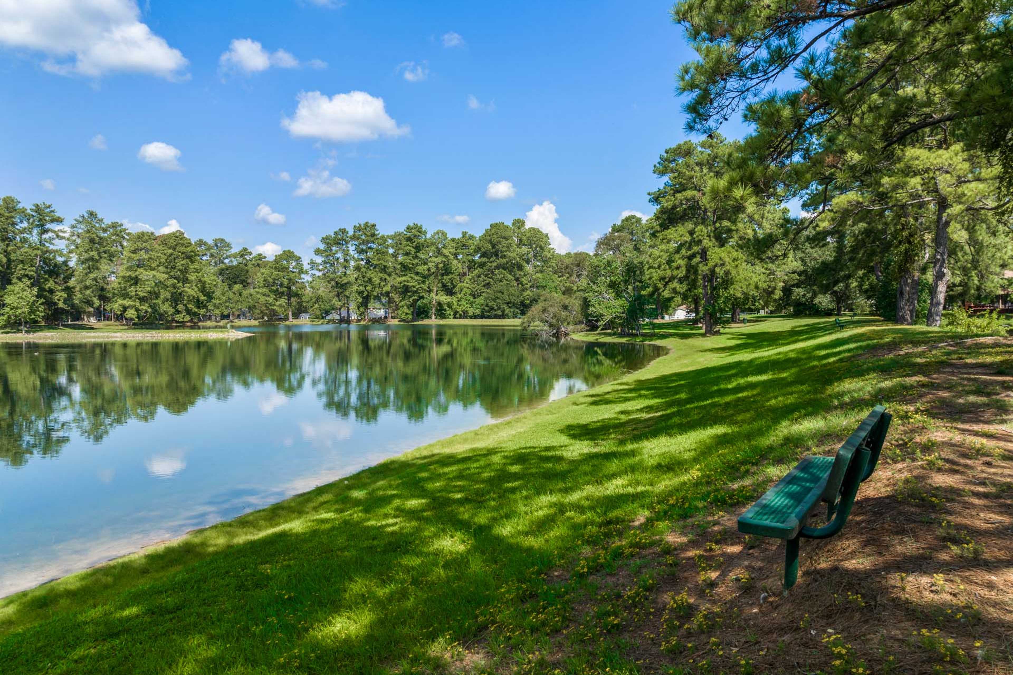 Serene pond reflecting pine trees and blue sky at Windmill Estates in Magnolia, Texas with green bench on grassy shore