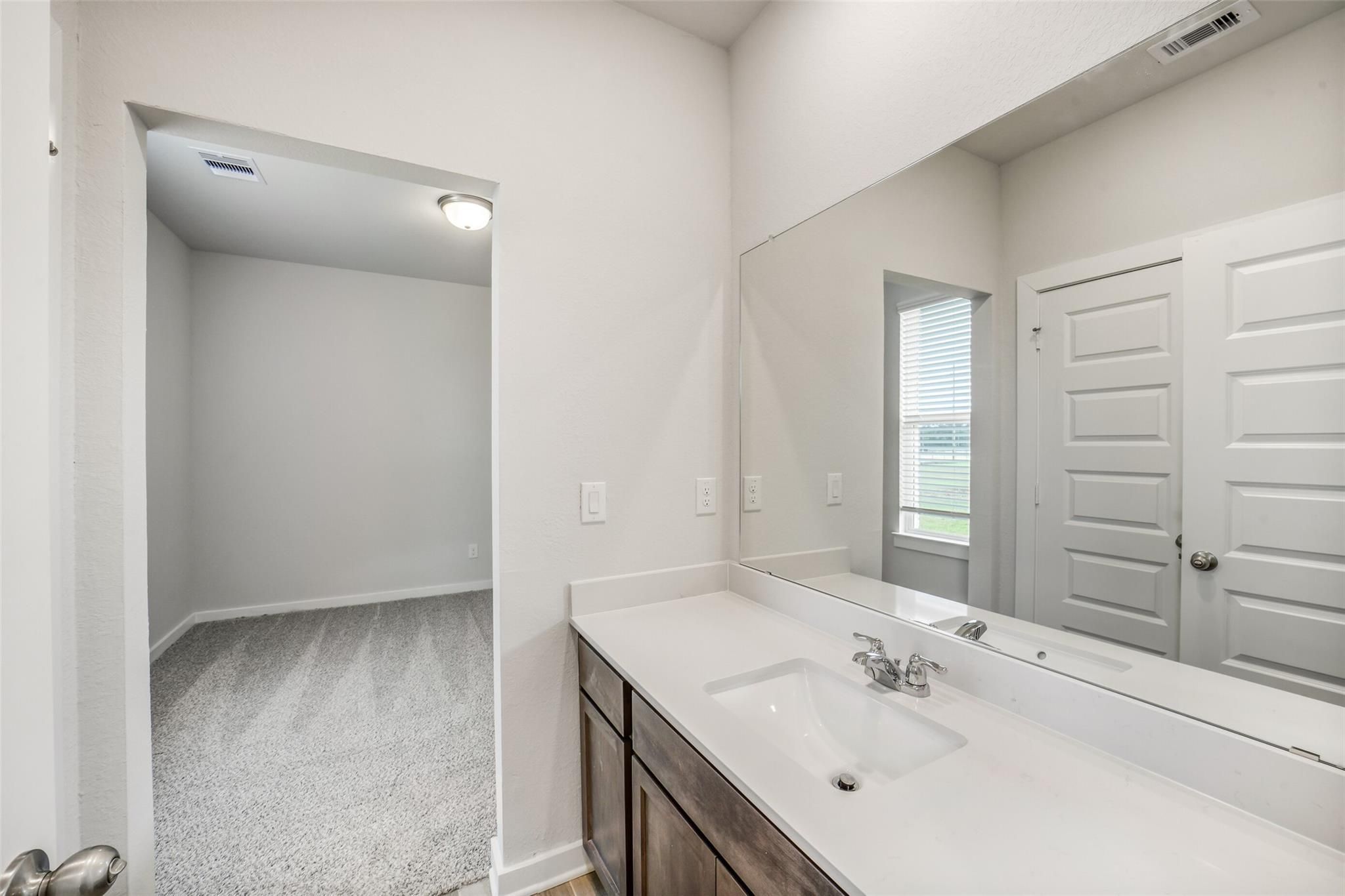 Modern bathroom vanity with white quartz countertop, single sink, large mirror, and adjacent carpeted closet in Davidson Homes The Acadia C, Magnolia, Texas