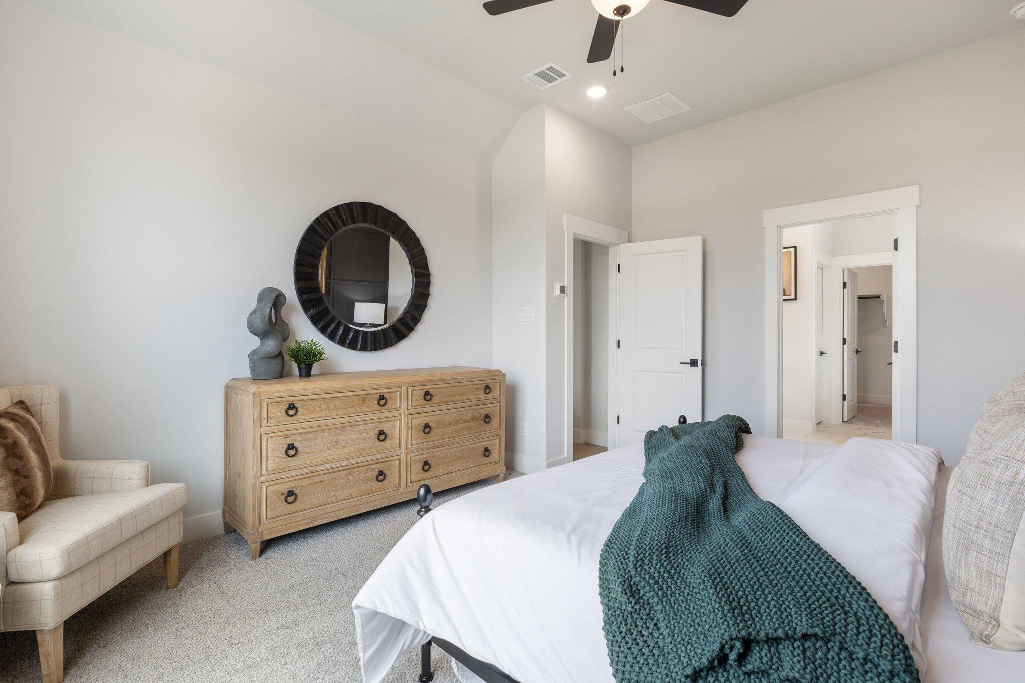 Master bedroom in The Sequoia M with white king bed, green throw, oak dresser, round black mirror, beige armchair, ceiling fan, walk-in closet