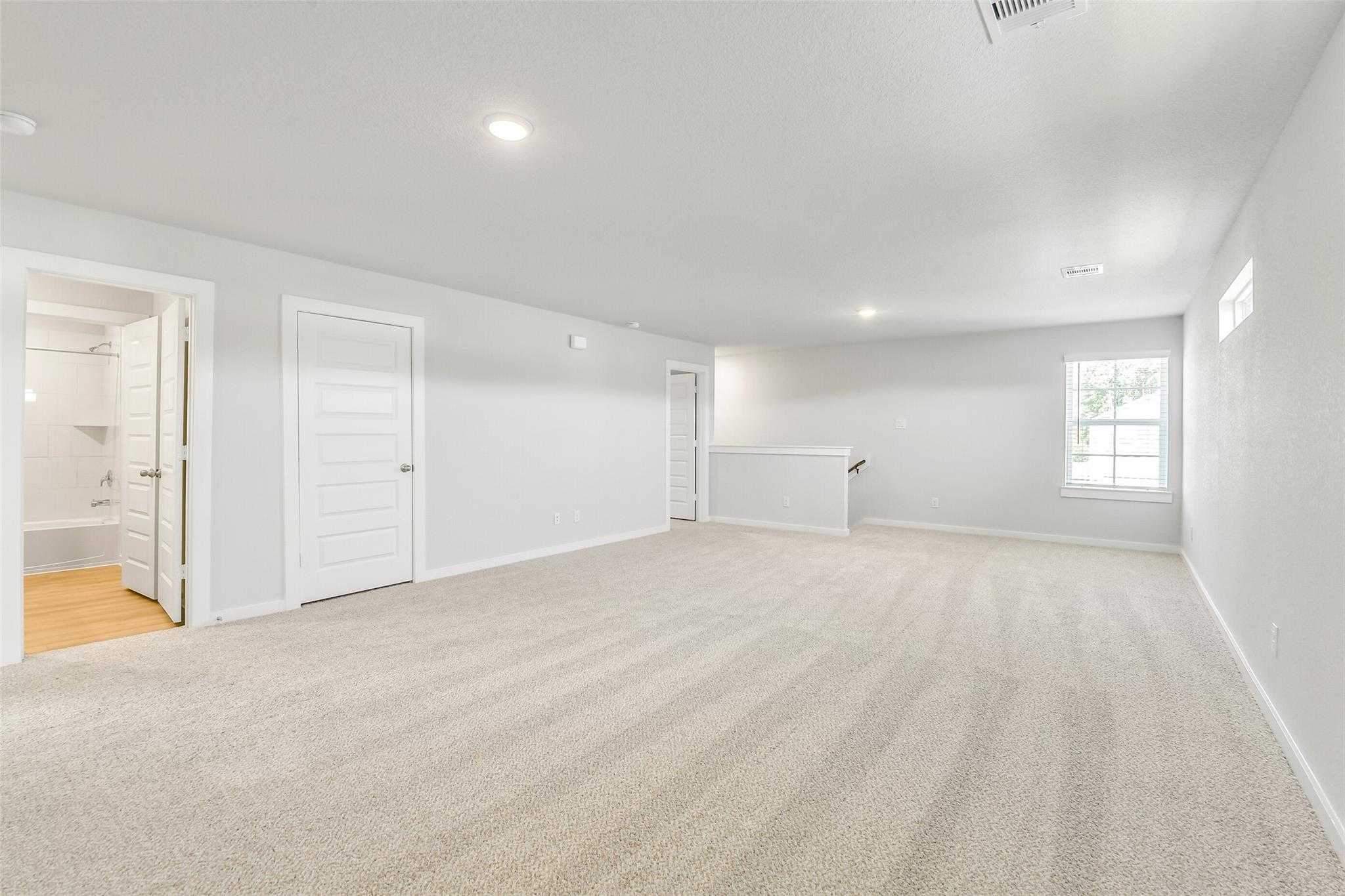 Spacious upstairs loft with beige carpet, white walls, adjacent bathroom, and window in 5-bedroom Davidson Homes The San Marcos E, Cleveland, Texas