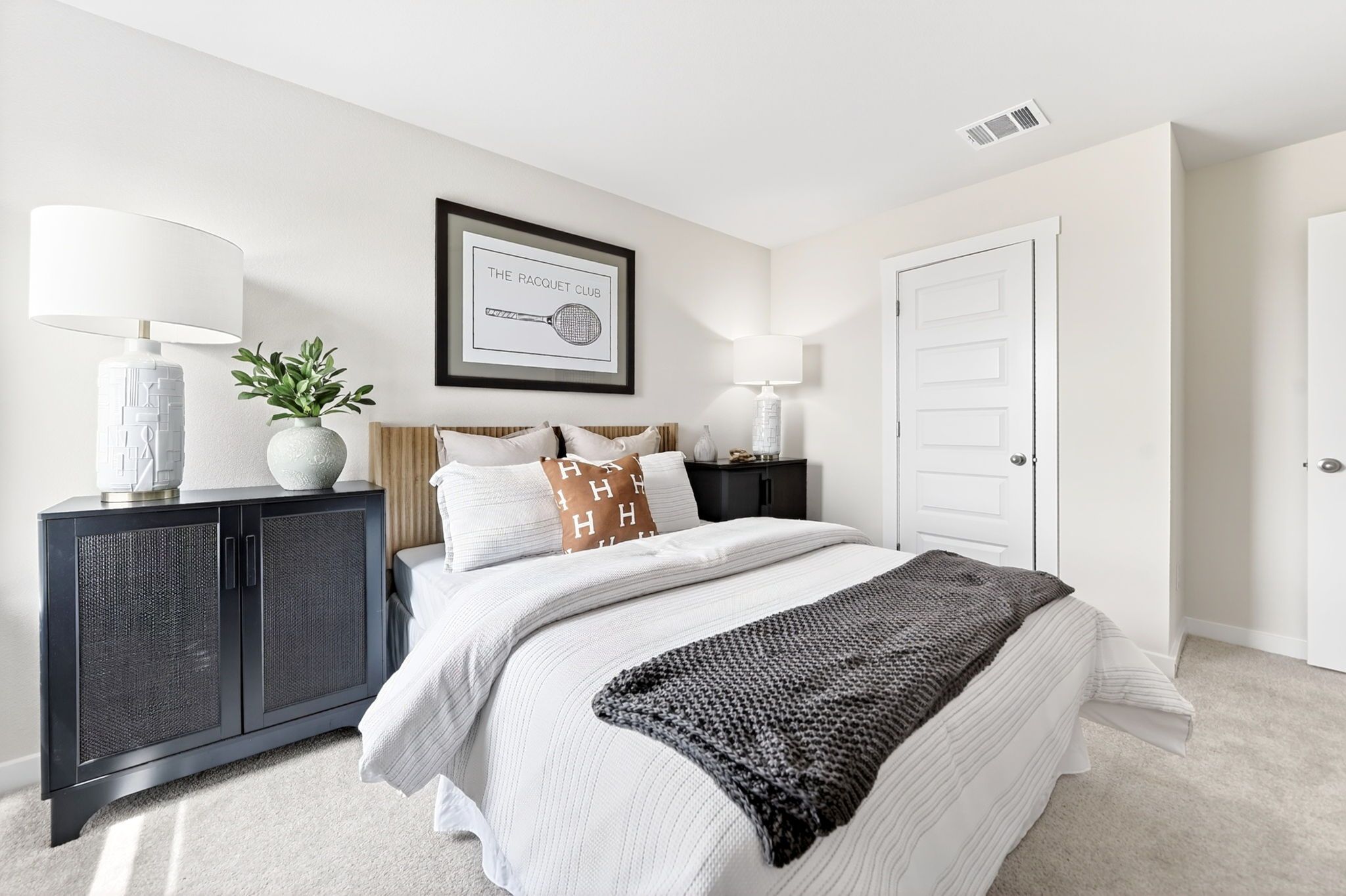 Cozy bedroom interior at Heartland Texas with white bedding, orange accent pillows, gray knit throw, rattan nightstands, and racquet club wall art