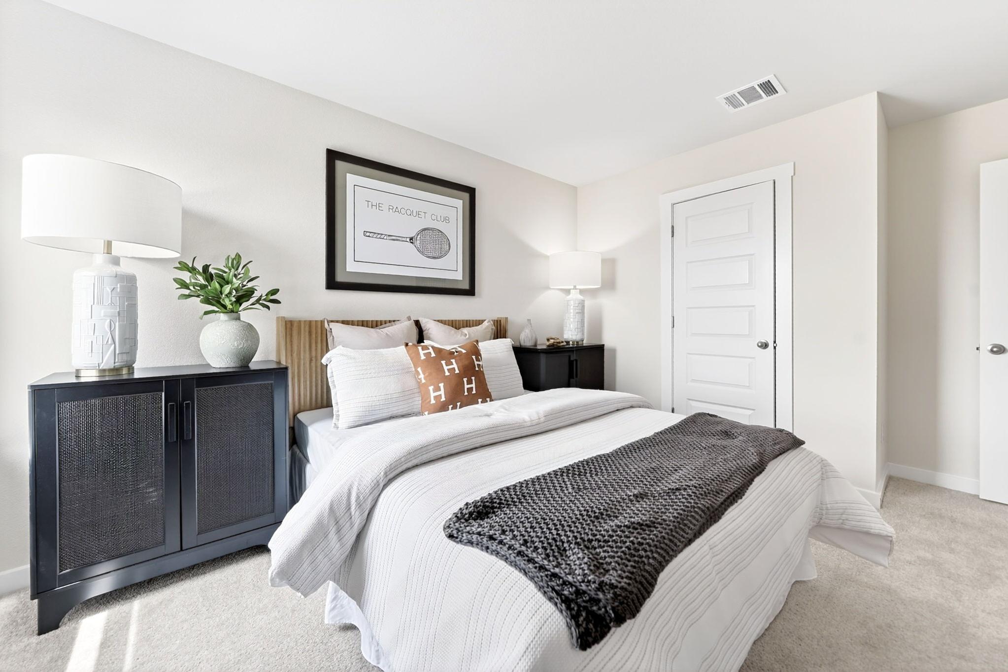 Cozy bedroom interior at Heartland Texas with white bedding, orange accent pillows, gray knit throw, rattan nightstands, and racquet club wall art